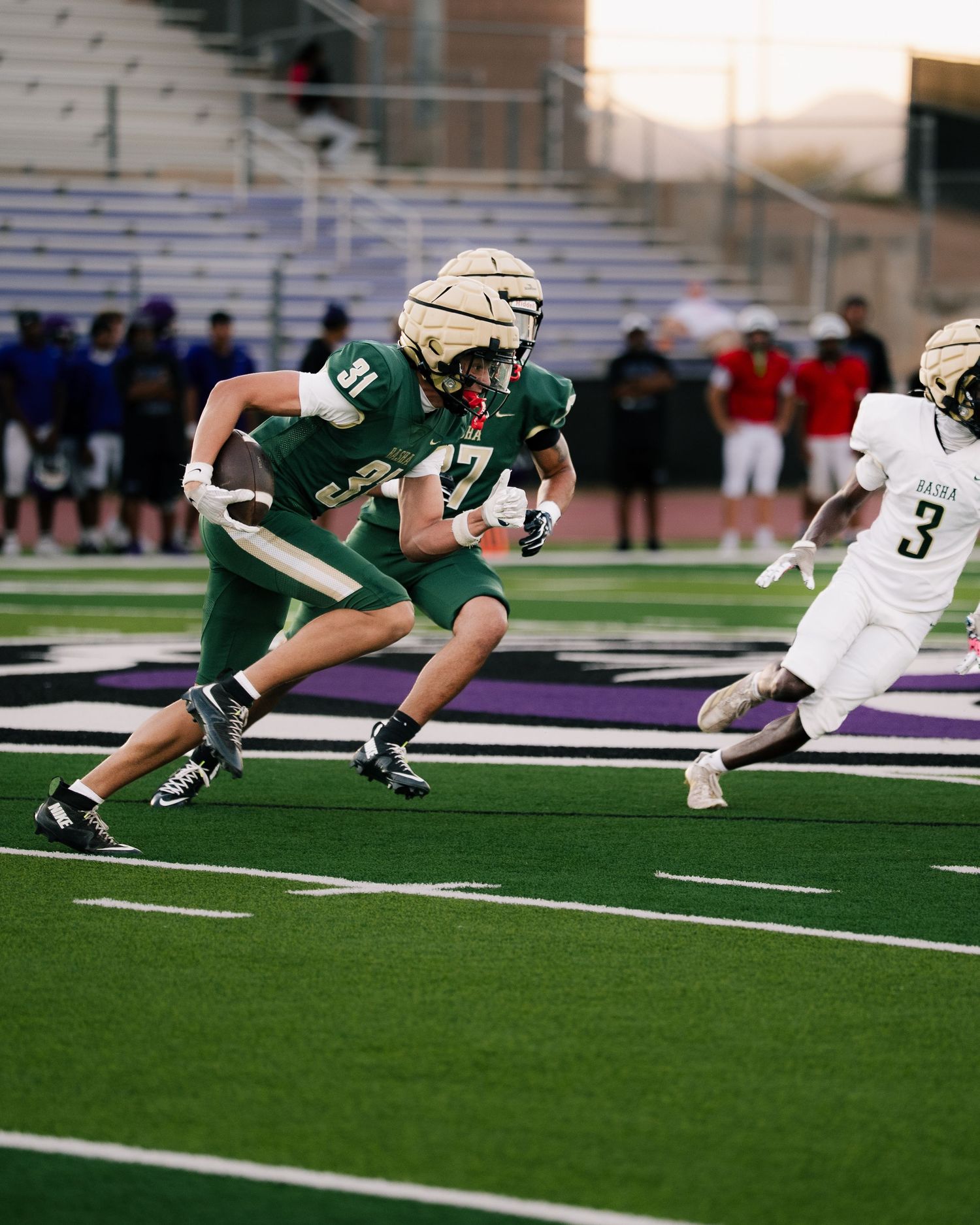 High school football player in green uniform runs with the ball as defender in white jersey number 3 pursues on turf field.