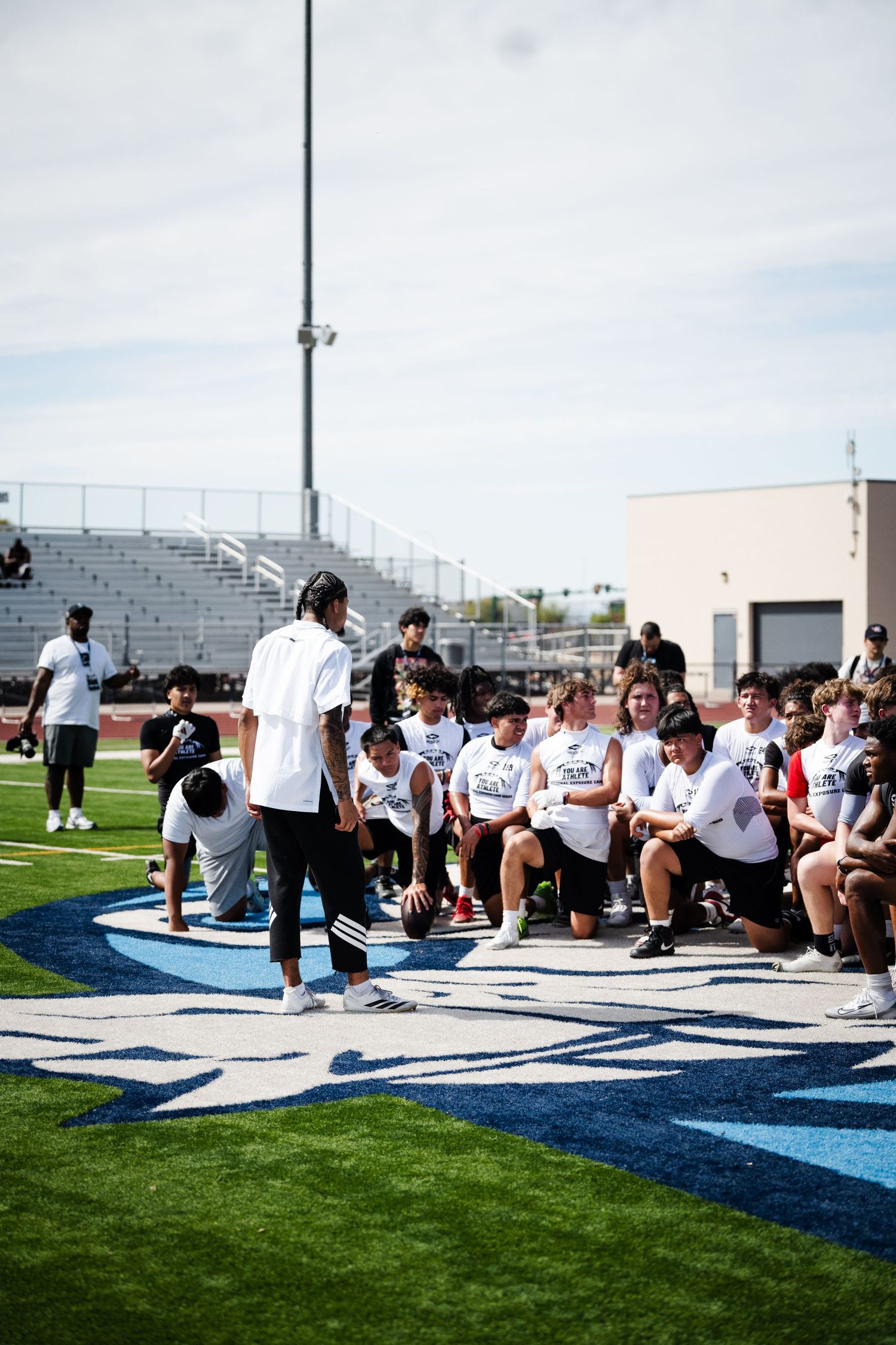 Coach addresses a group of young football players seated on a turf field during a training camp.
