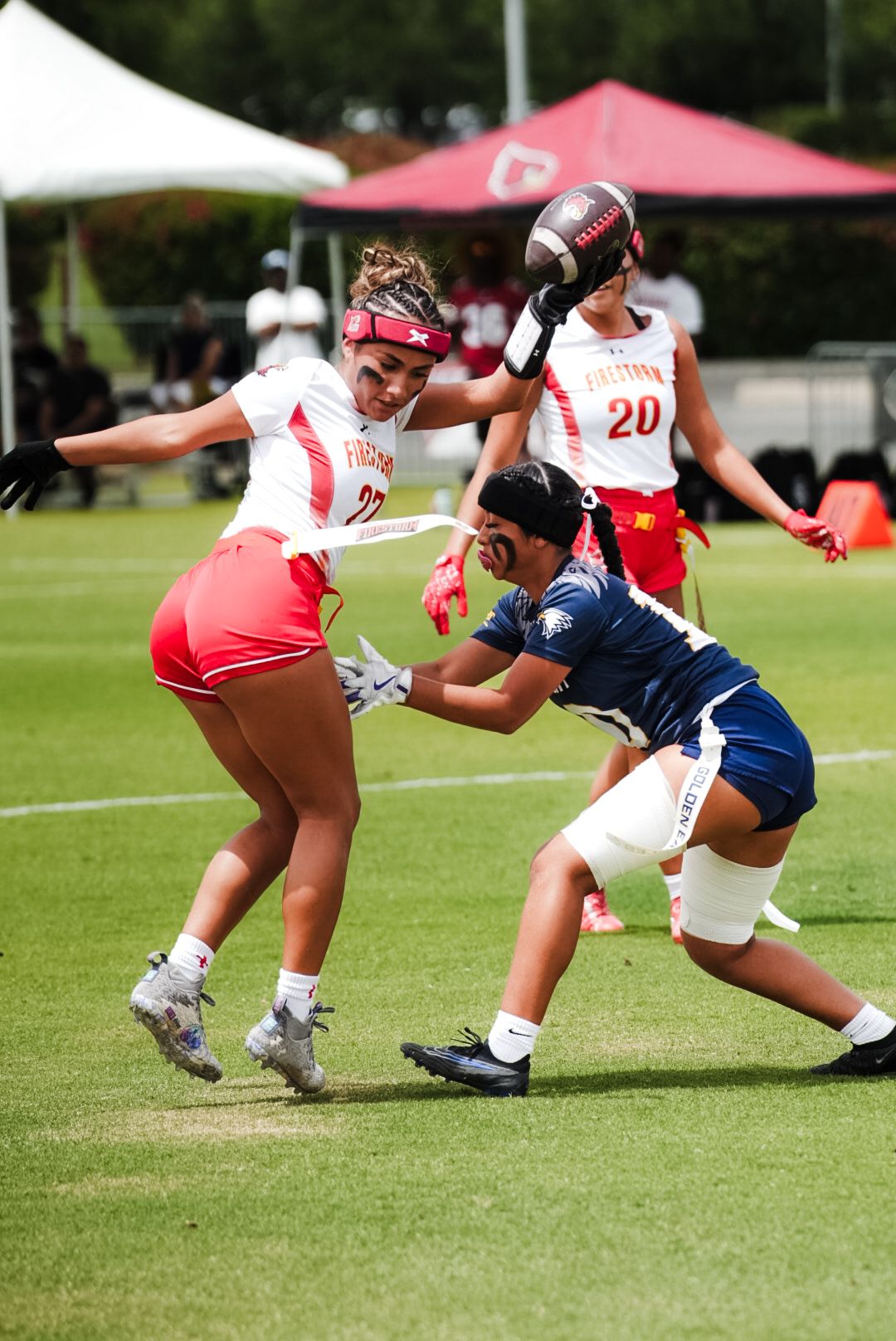 Women playing flag football, player in red jersey attempts to grab flag from opponent in navy uniform on green field.
