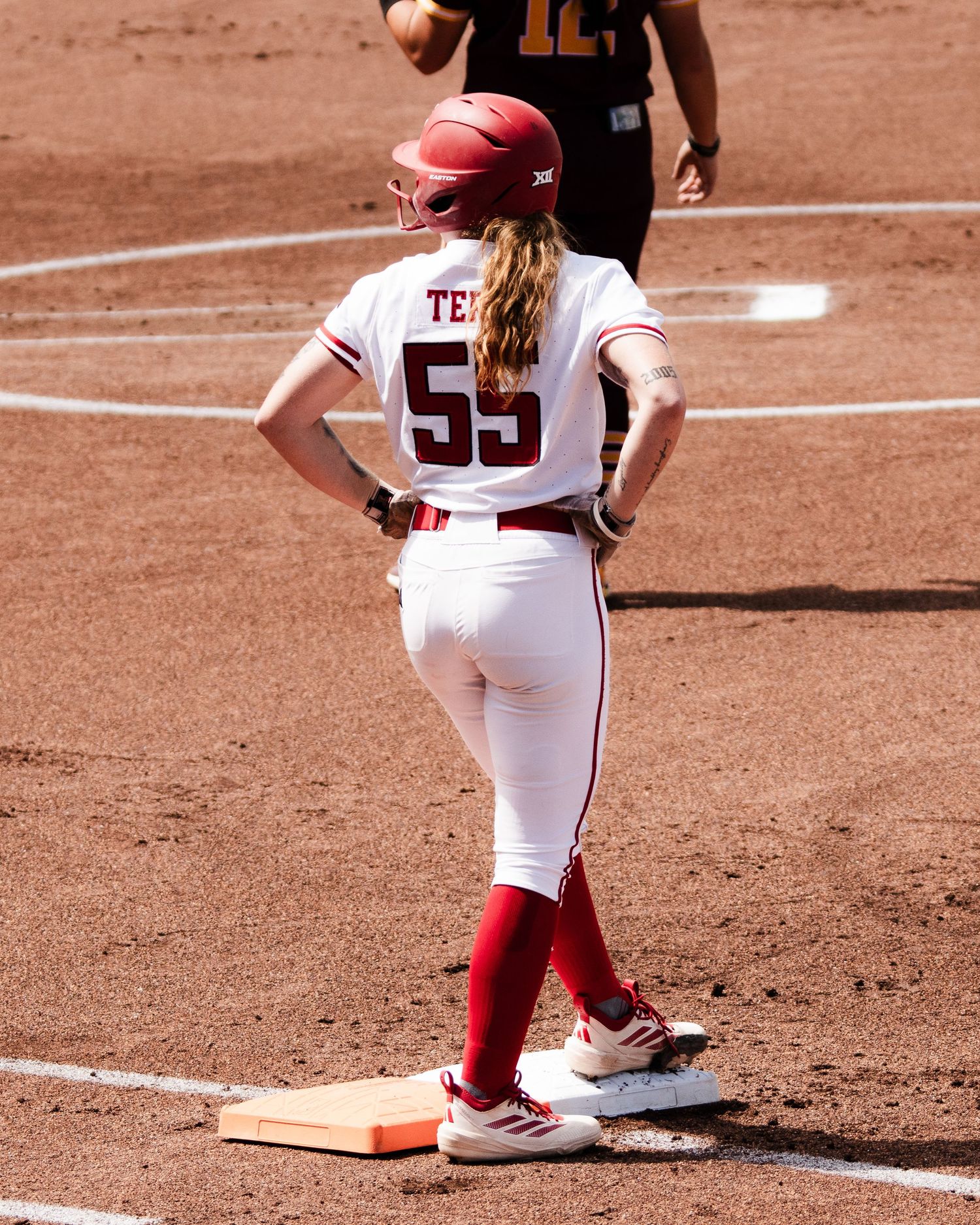 Softball player number 55 in white uniform standing on base during college game.