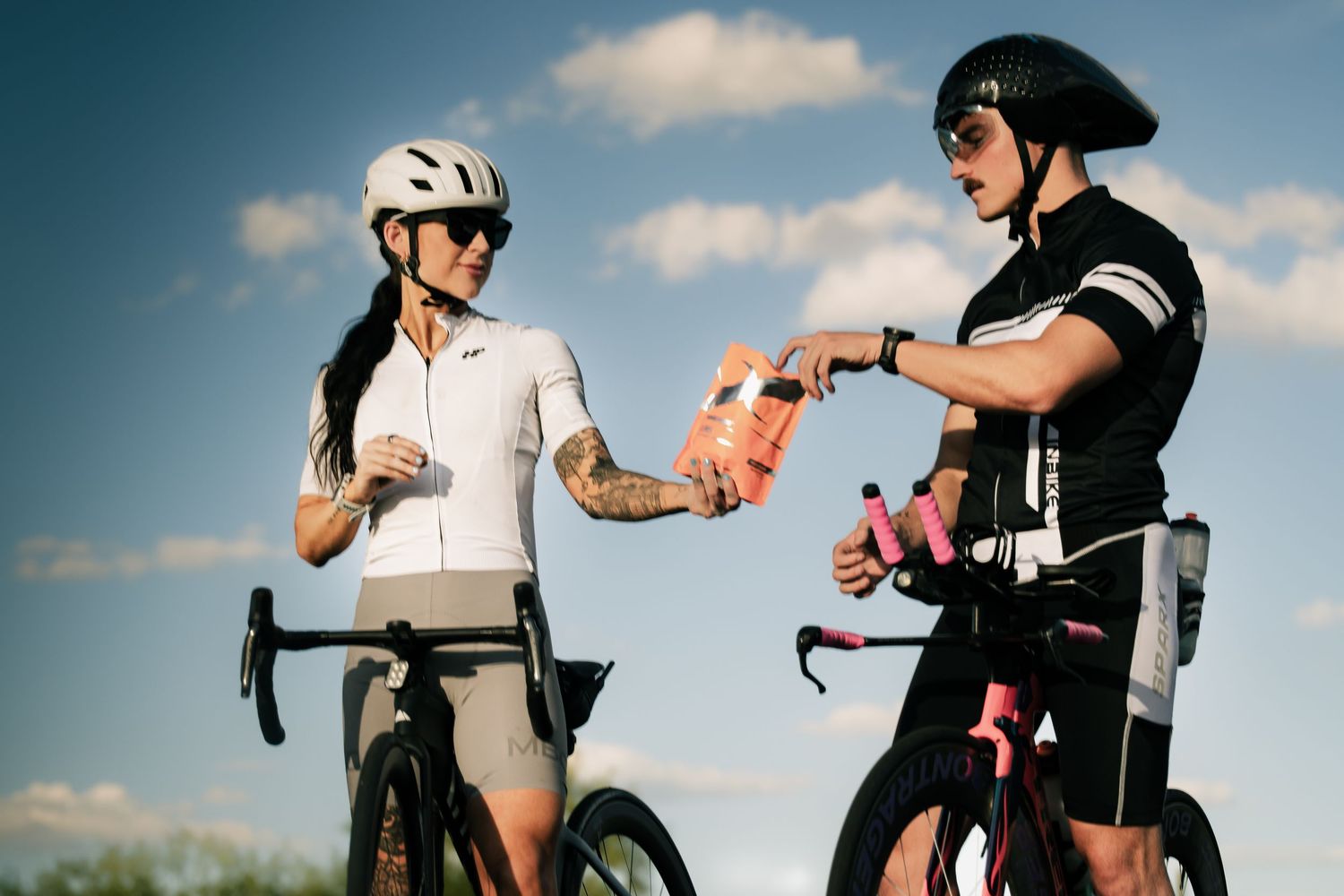 Two cyclists in helmets pause outdoors, sharing a pink water bottle against a blue sky backdrop.