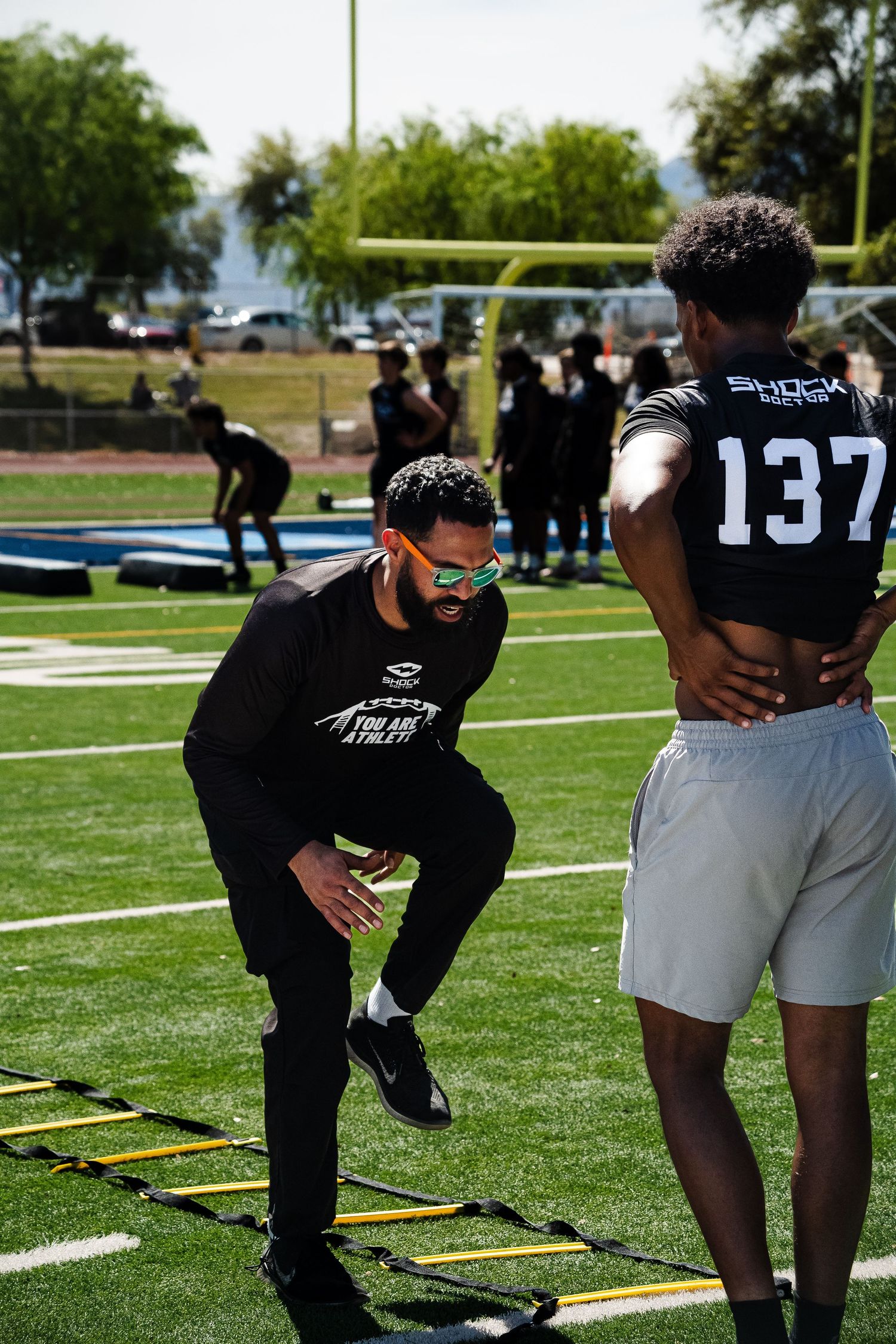 Football coach in black demonstrates agility ladder drill to player #137 on green turf field.