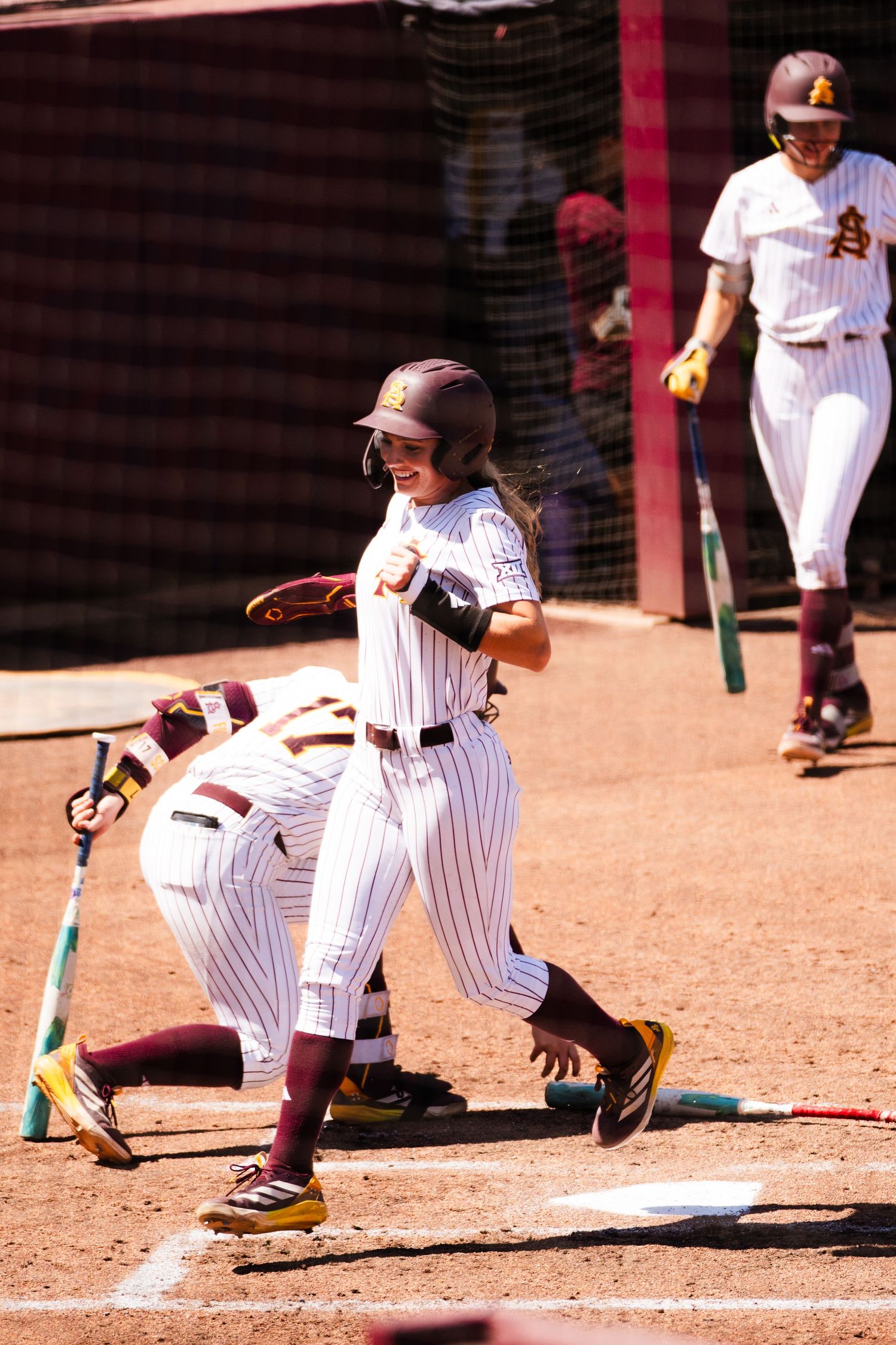 College softball player smiling at home plate in maroon pinstripe uniform, with teammate in background.