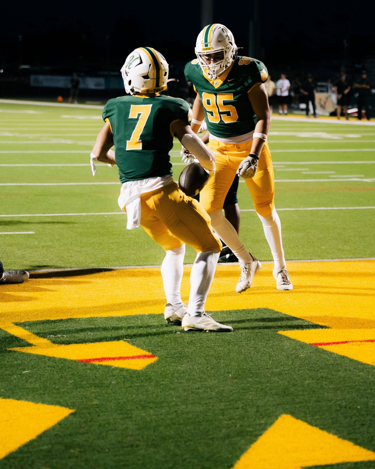 Football players #7 and #95 in green and gold uniforms celebrate on the field during a night game.