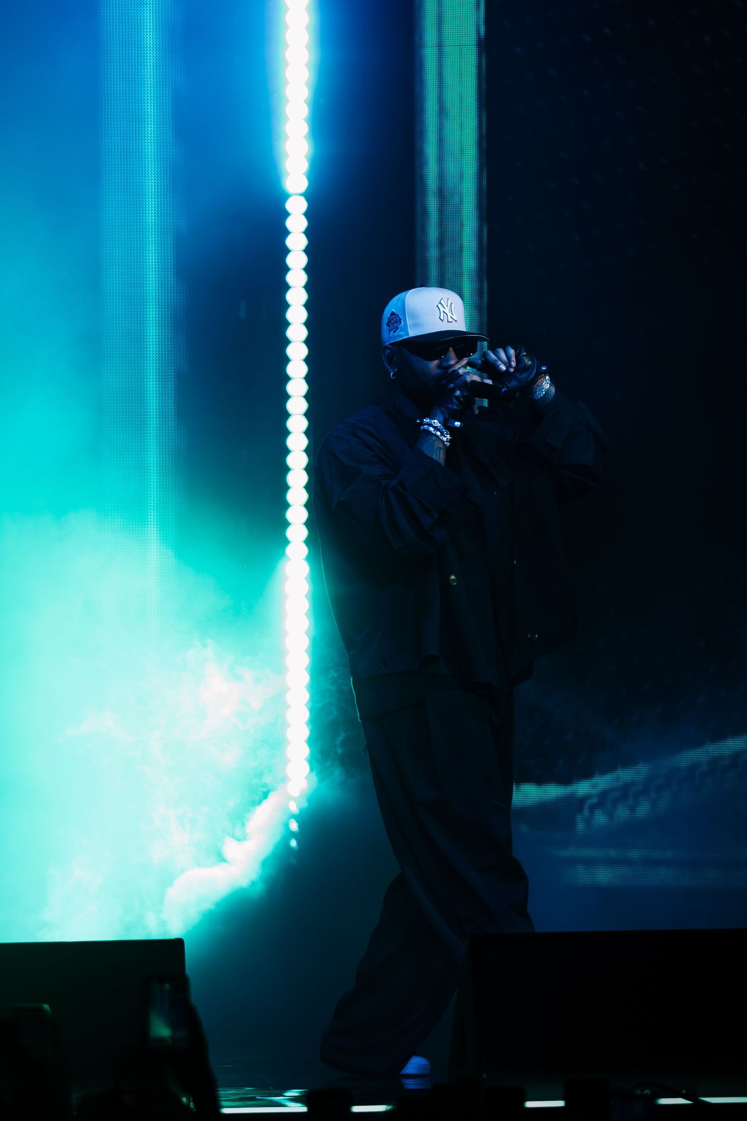 Performer in a white cap and dark clothing holds a microphone on stage, lit by glowing teal lights and haze during a live concert.