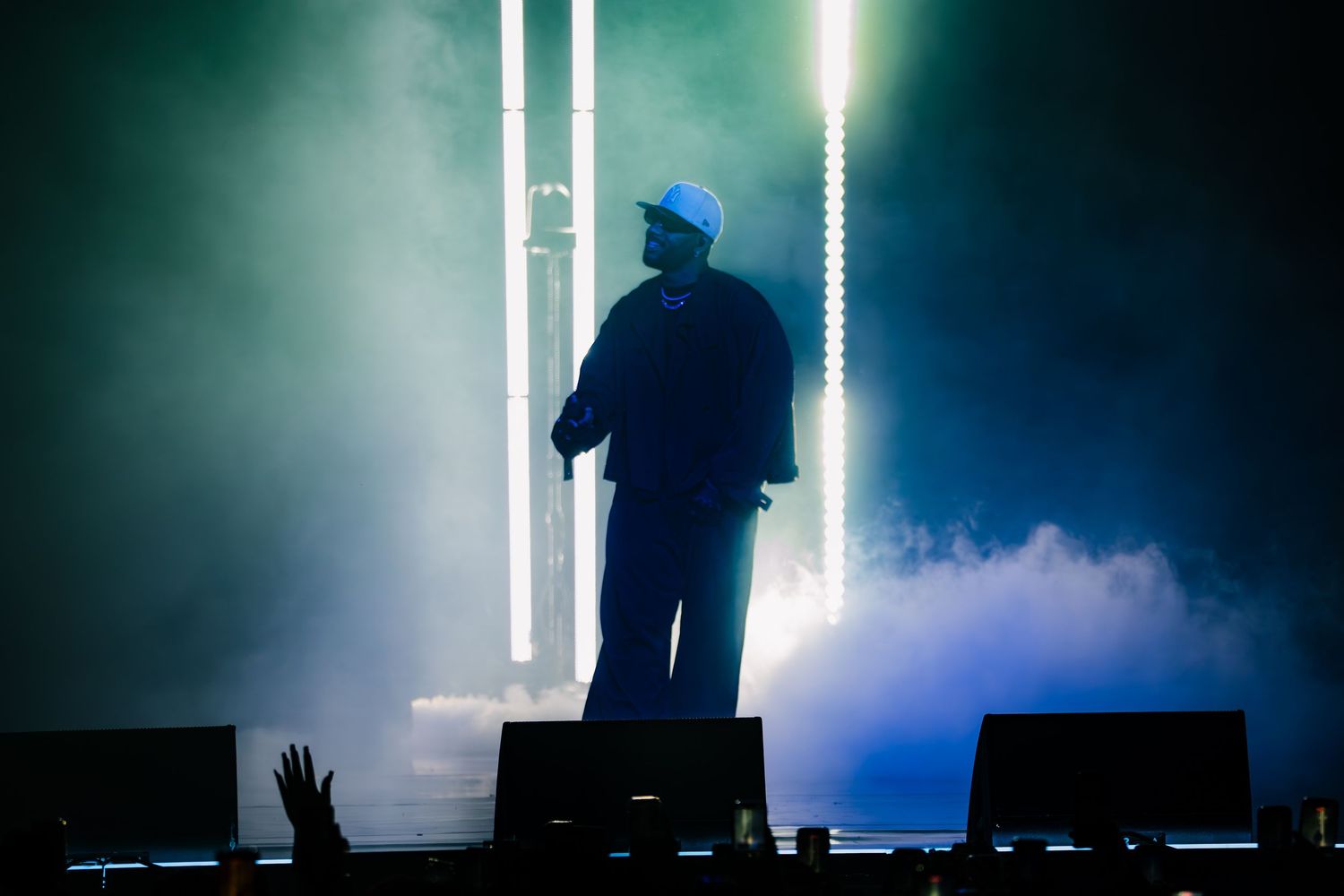 Silhouetted performer in a light cap stands on a smoky stage with glowing vertical light bars while a fan's raised hand appears in the foreg