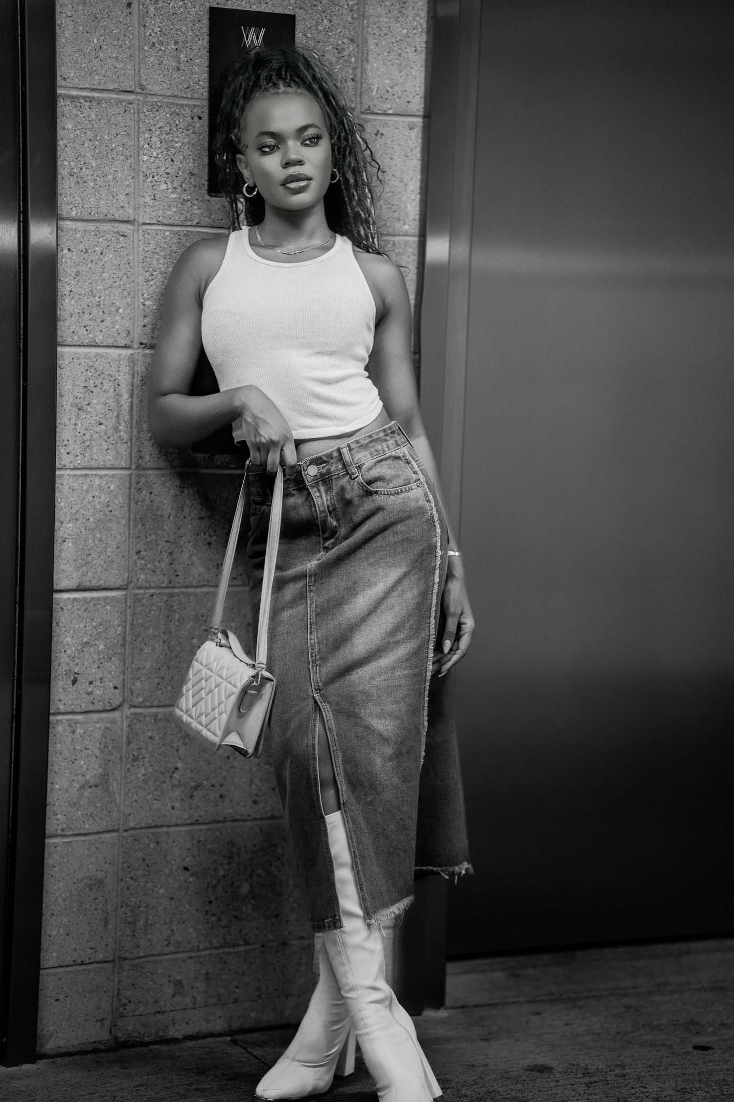 Black and white photo of a stylish woman in white tank top, denim skirt, and white boots leaning against a wall.
