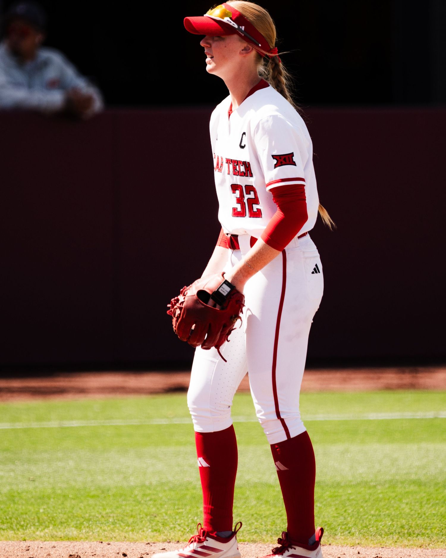 Female softball player #32 in red and white uniform holds glove on sunny field.
