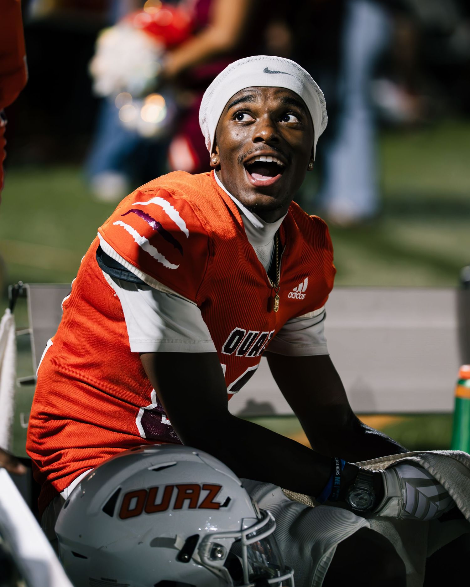A football player in an orange Adidas jersey sits on the sideline smiling, wearing a white Nike cap and holding a helmet.