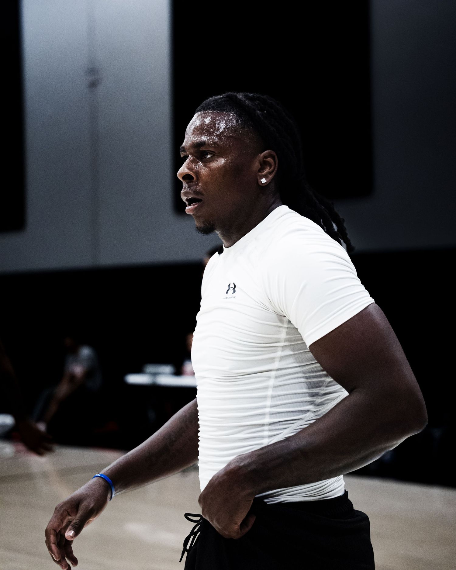 Athletic man in white Under Armour compression shirt with braided hair stands on basketball court.