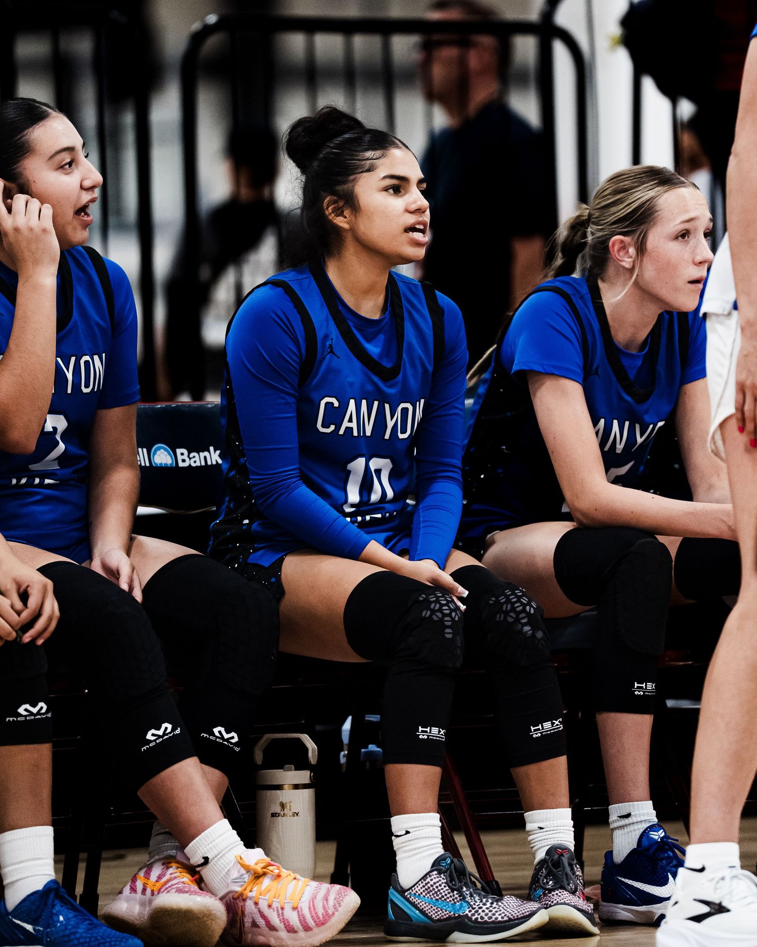 Three female Canyon basketball players in blue uniforms sit attentively on bench during game.