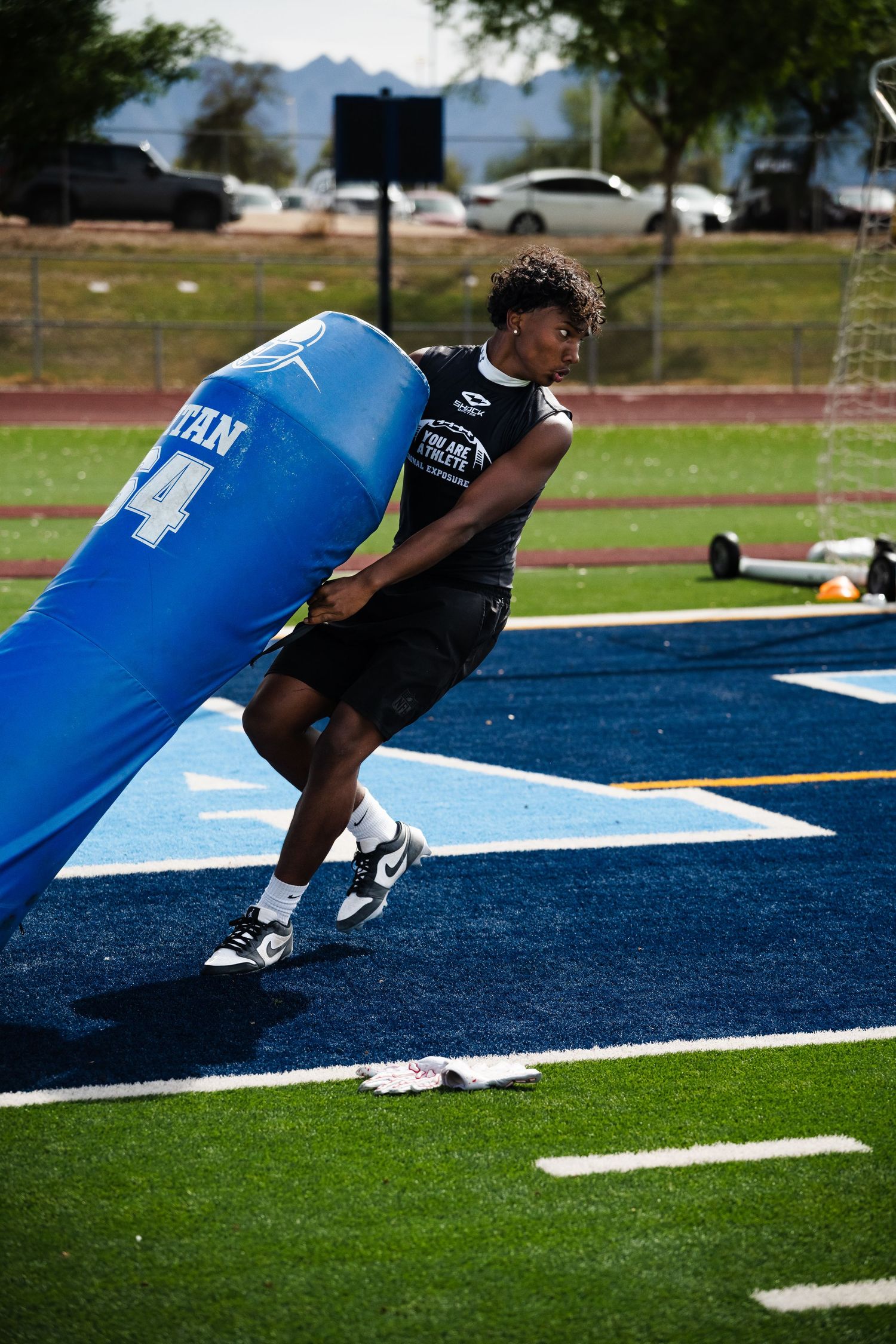 Football player drives into blue blocking dummy during defensive line drill on turf field.