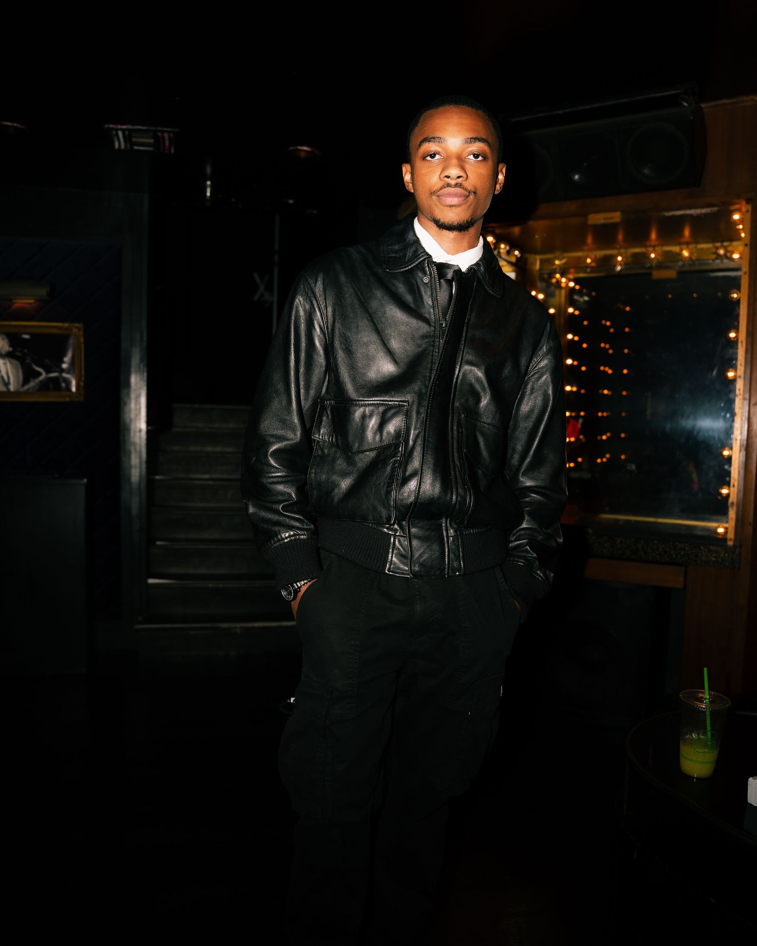 Young man in black leather jacket and pants posing indoors at a dimly lit venue with warm string lights.