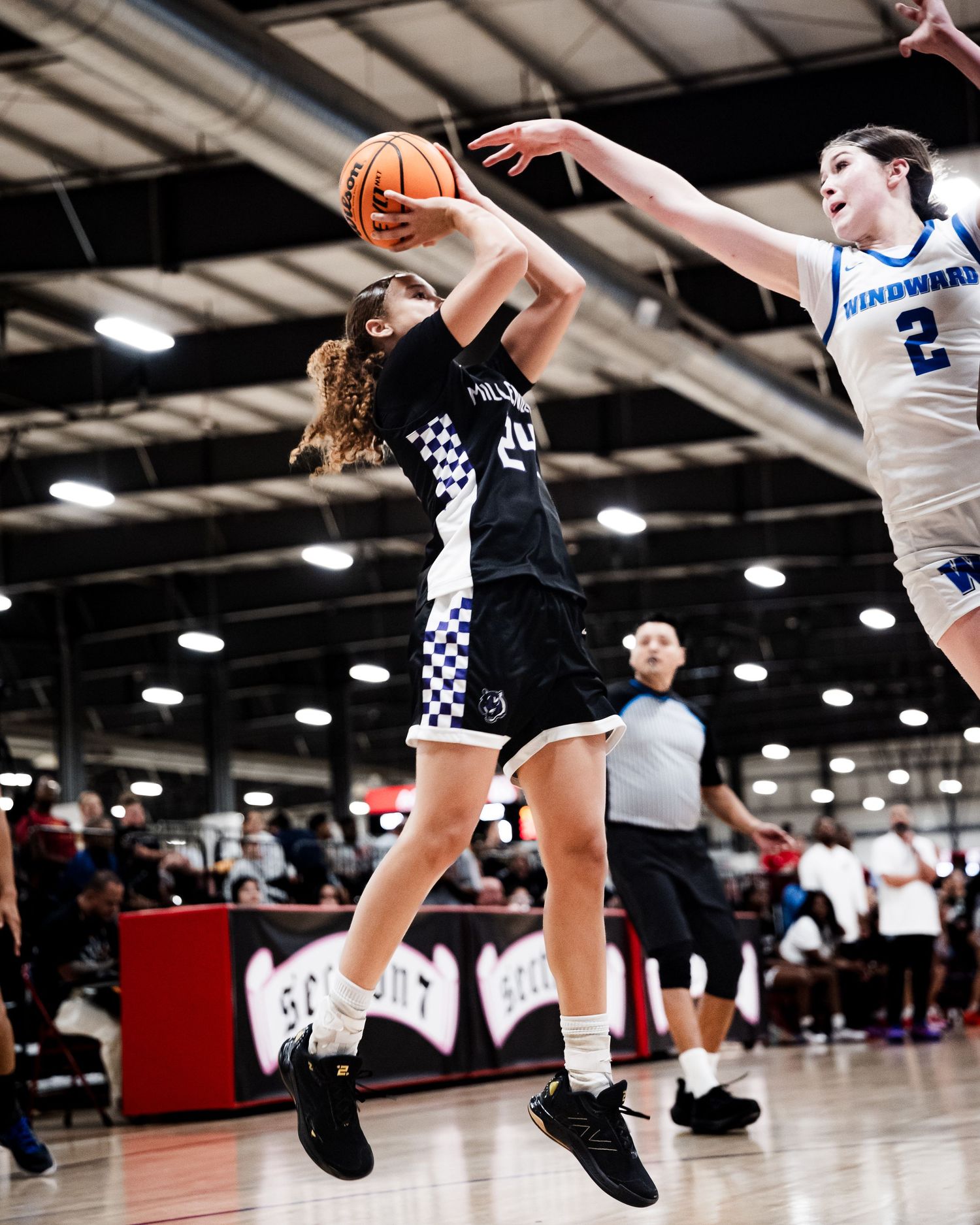 Female basketball player in black Millennium uniform drives to basket against Windward defender indoors.