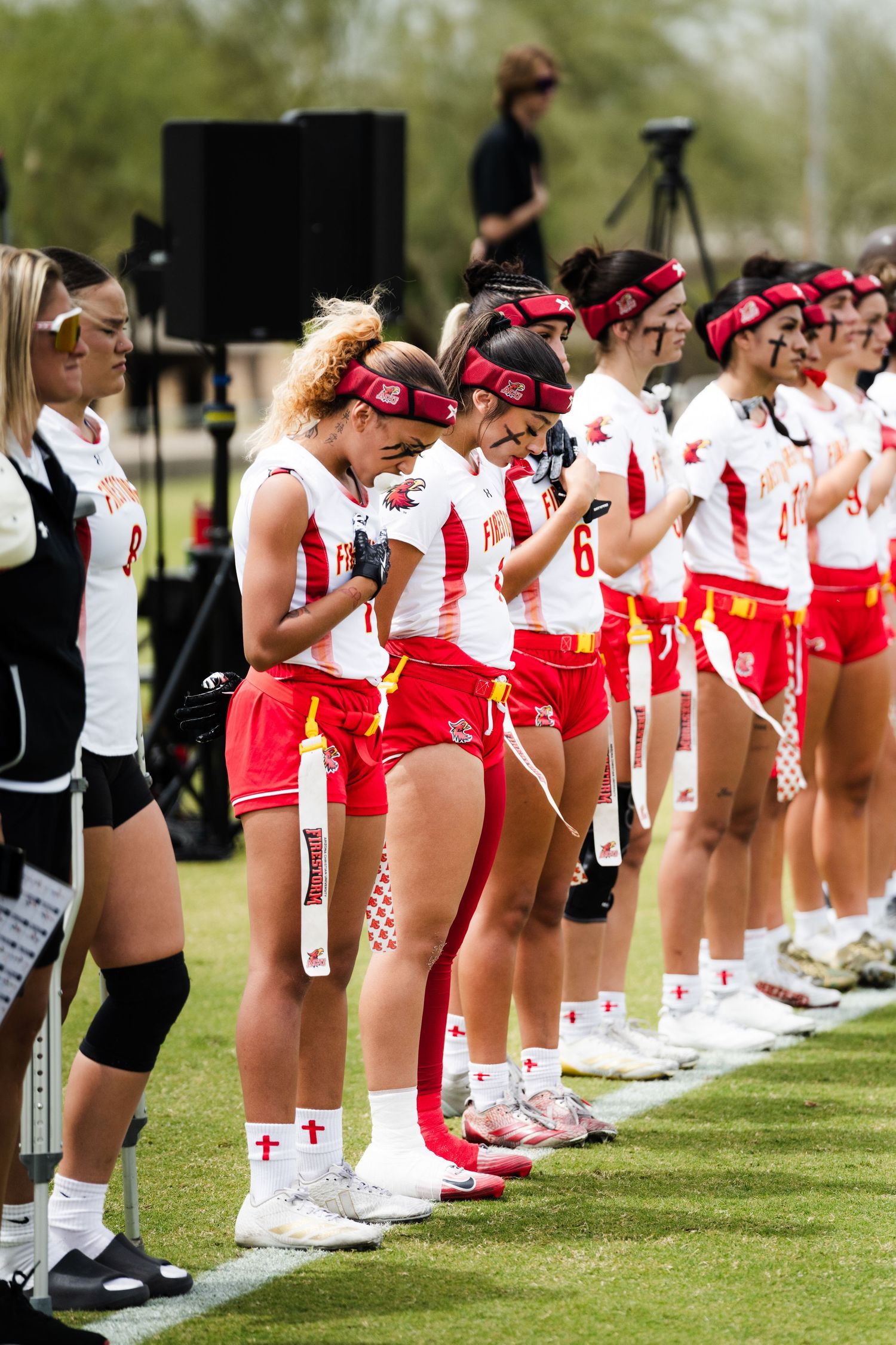 Female flag football team in red and white uniforms lines up on the sideline during a pre-game ceremony outdoors.