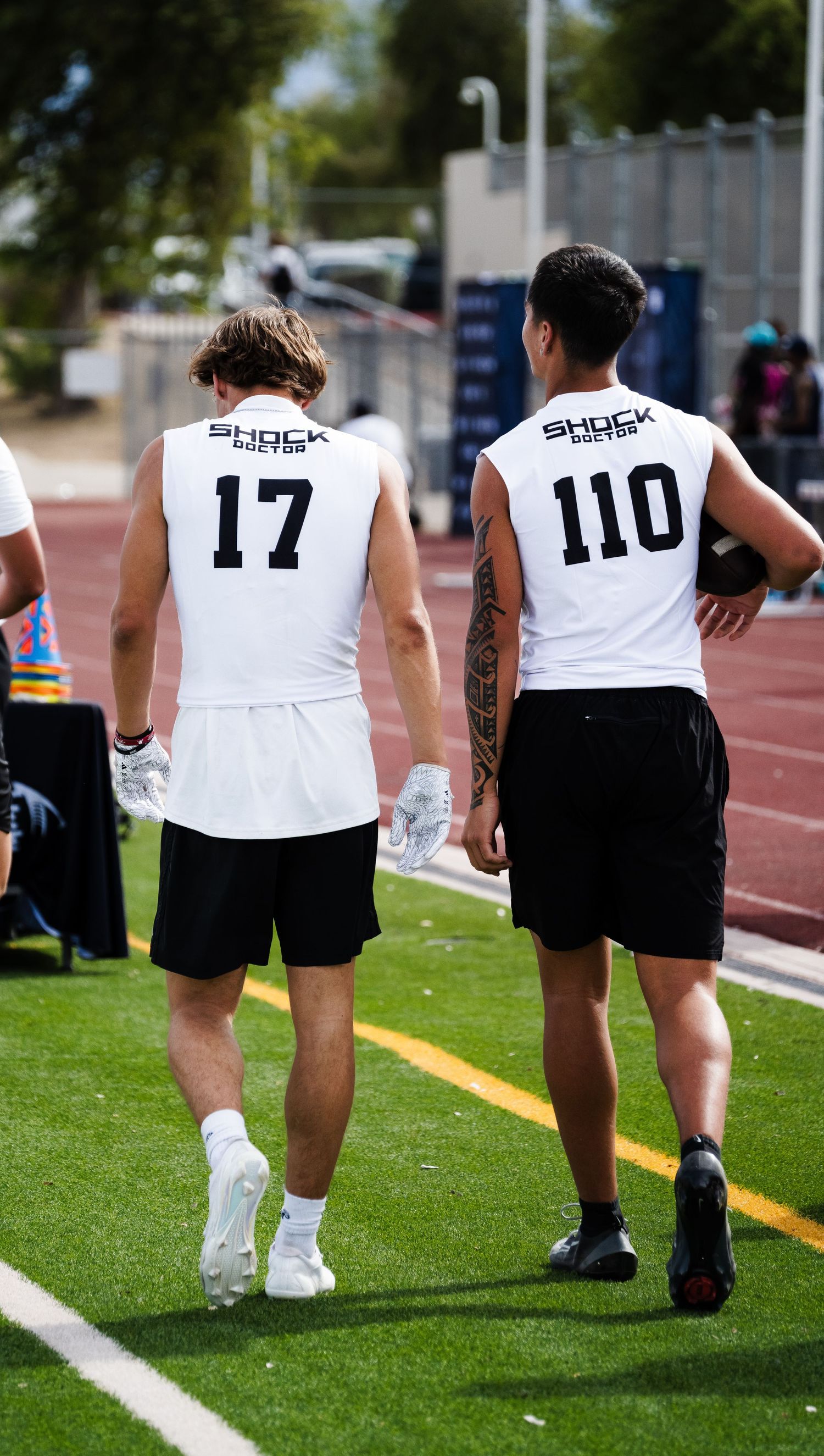 Two football recruits wearing Shock Doctor jerseys numbered 17 and 110 walking on sideline.