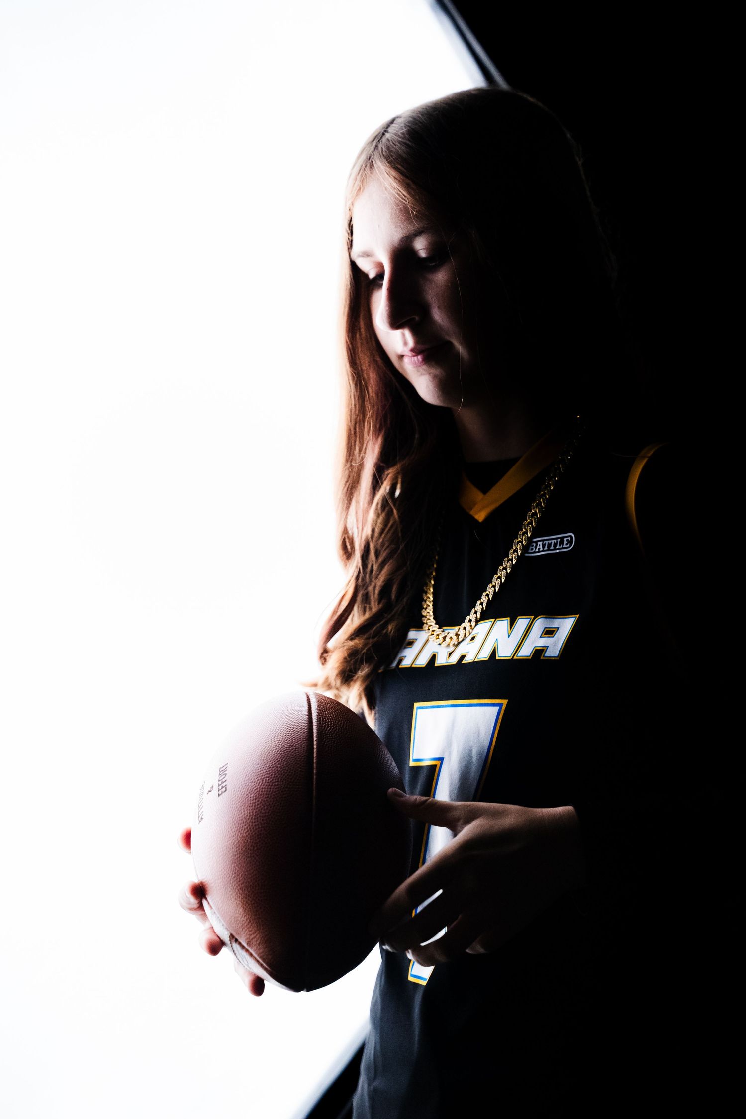 Female football player in dark jersey holding a football, dramatic backlit silhouette portrait with gold chain necklace.