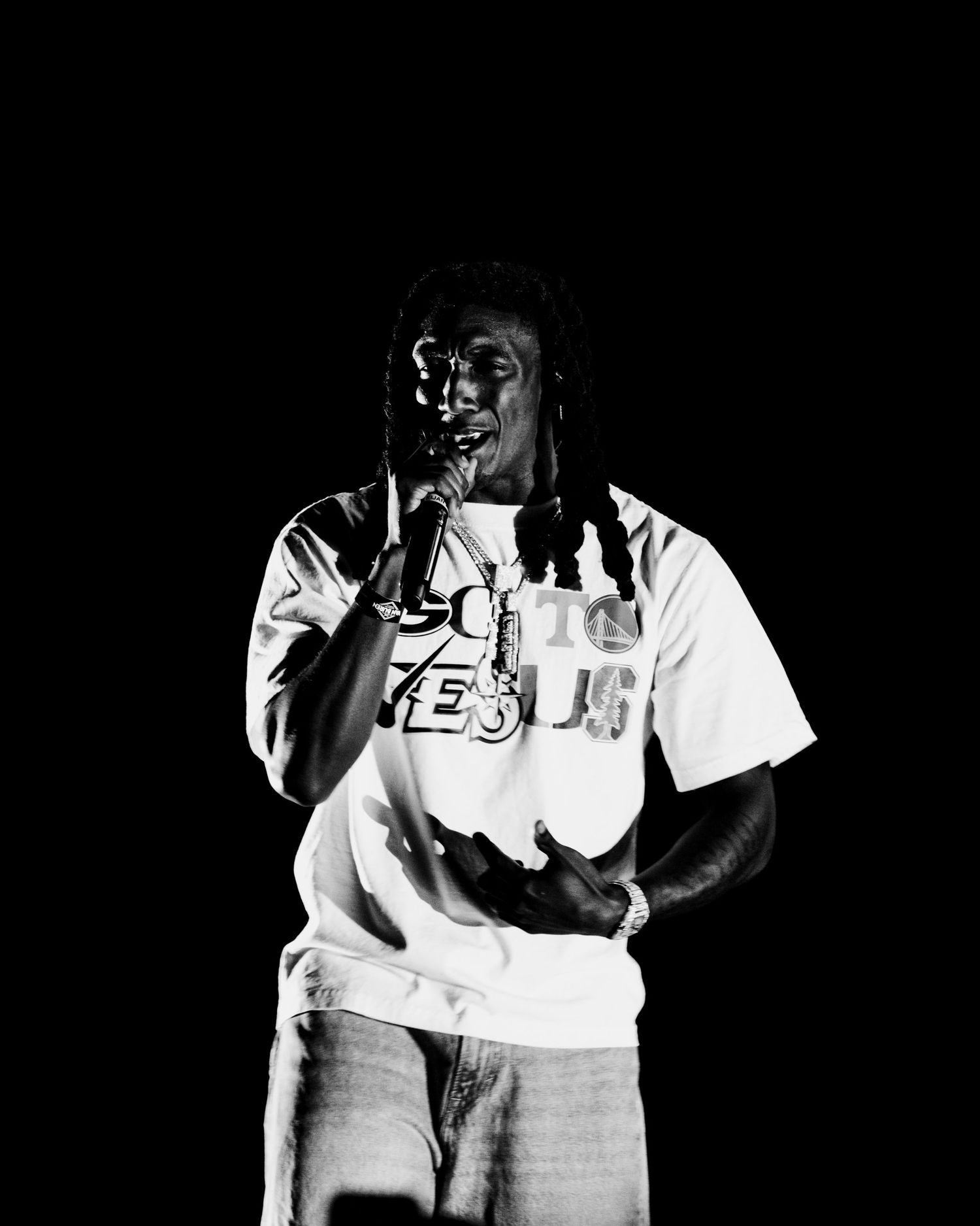 Black and white portrait of dreadlocked rapper performing with microphone on dark concert stage.