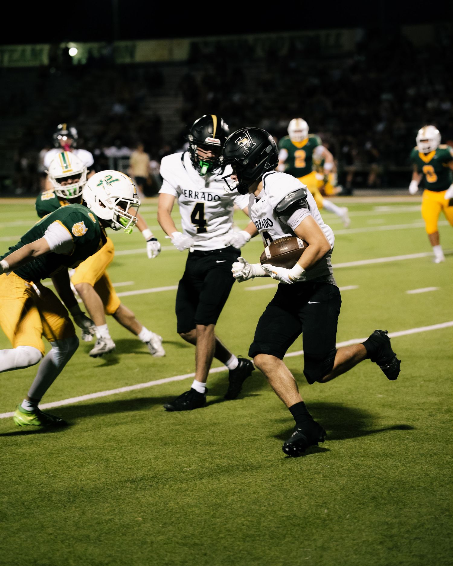 Football player in black uniform runs with ball while teammate blocks defender during night game.