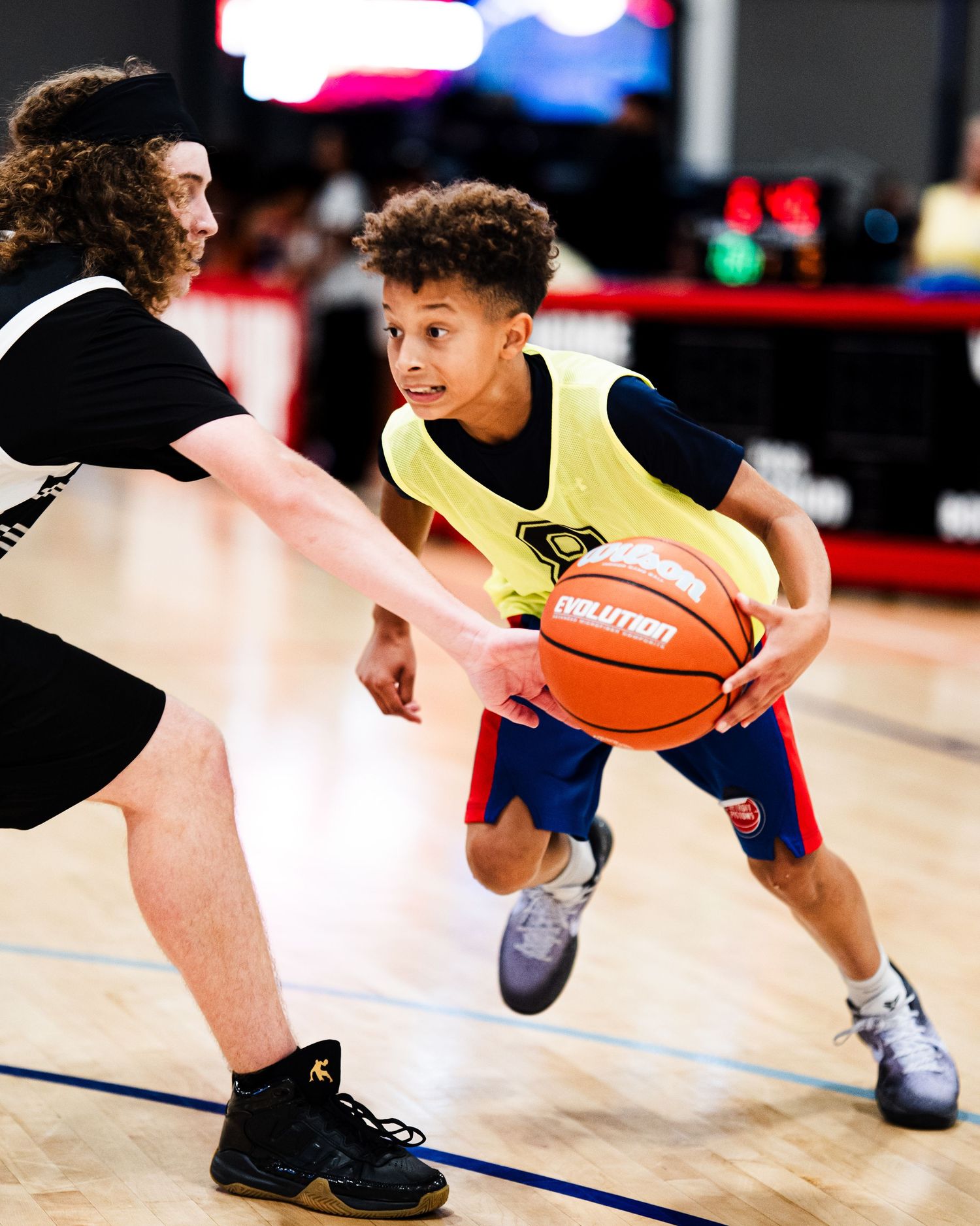 Young basketball player in yellow vest dribbles past defender during indoor youth basketball game.