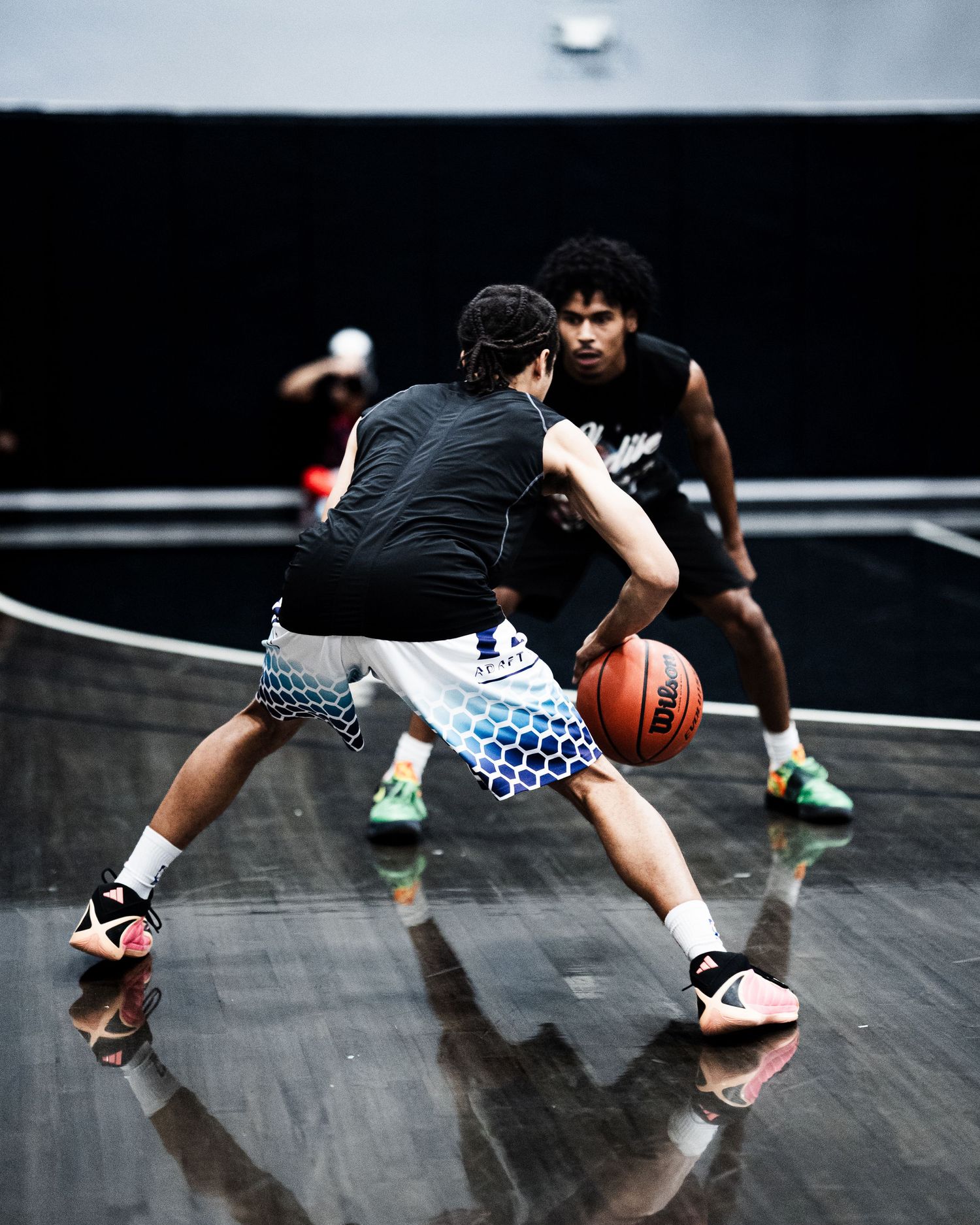 Two basketball players practice dribbling and defense on a shiny indoor court under dramatic lighting.