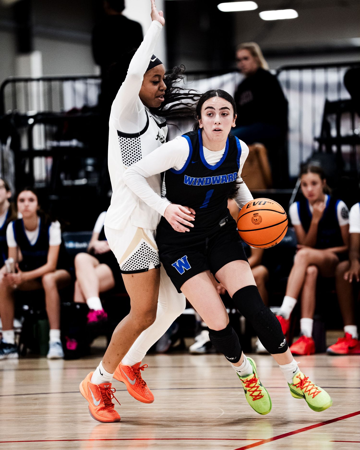 Female basketball player in blue Windward uniform drives to basket against defender during indoor game.