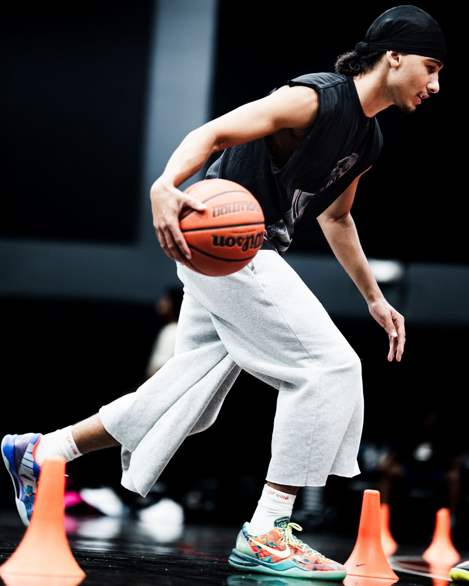 Player dribbling a Wilson basketball through orange cones during practice wearing gray sweats and colorful sneakers.