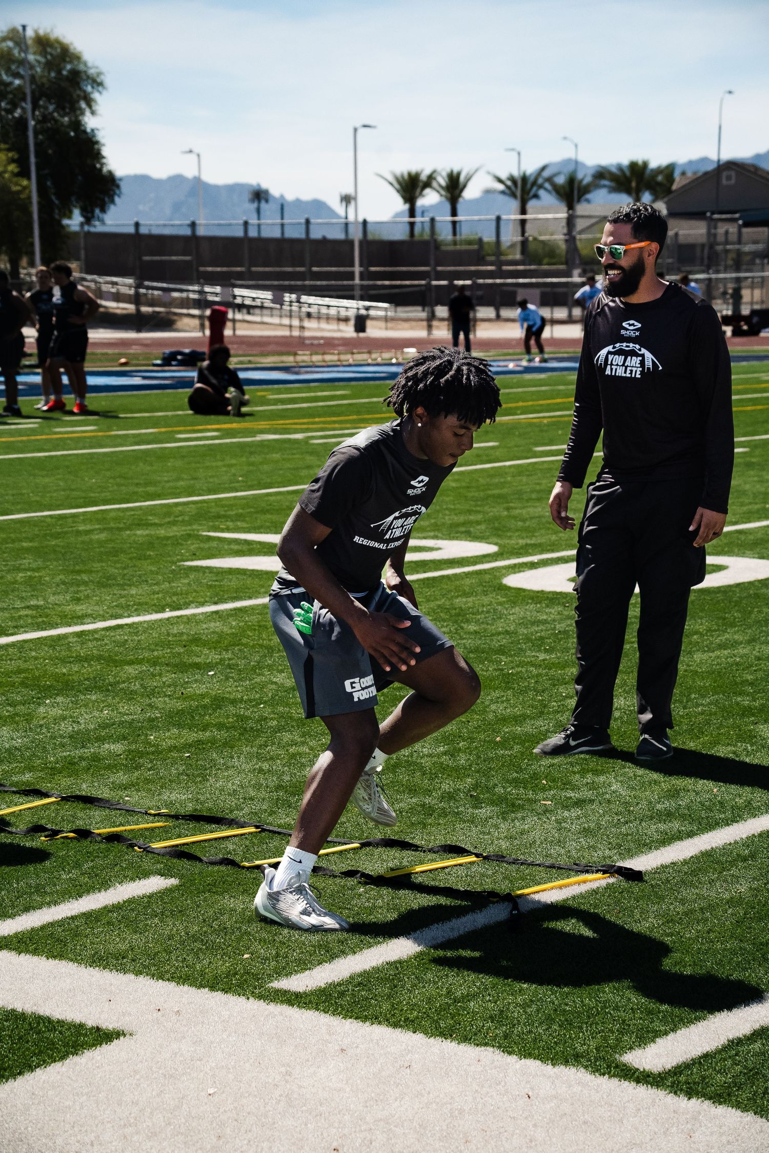Football coach observes young athlete completing speed ladder drill on outdoor turf field.
