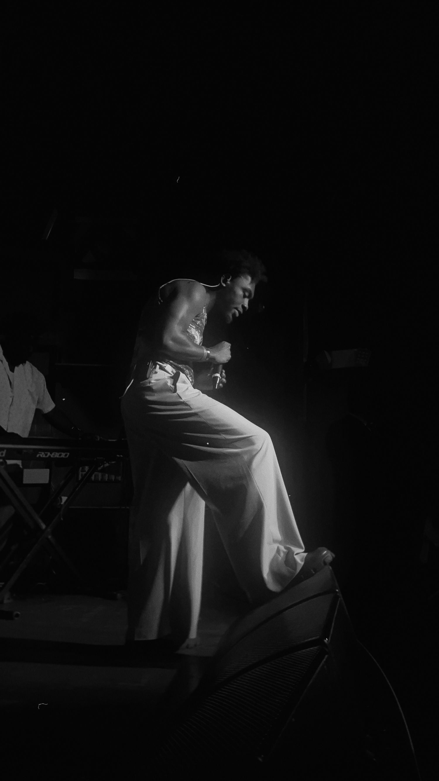 Black and white photo of a performer in wide-leg pants leaning forward on stage under dramatic spotlight.