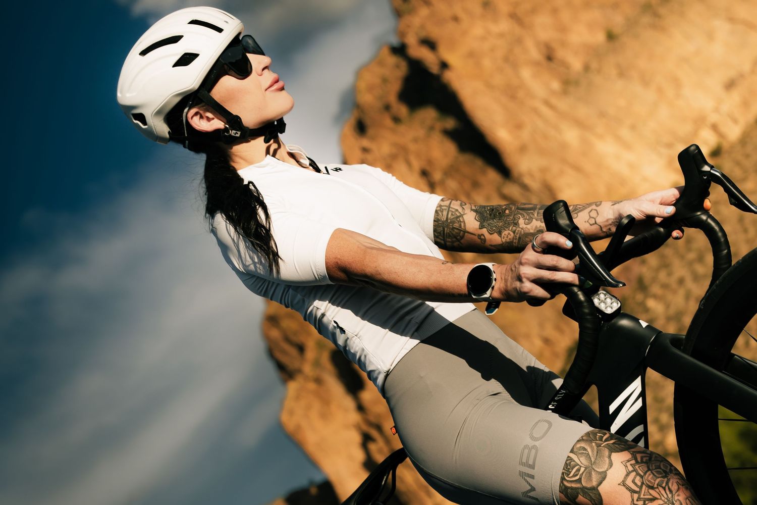 Tattooed female cyclist in white helmet and jersey poses with bike against rocky terrain under blue sky.