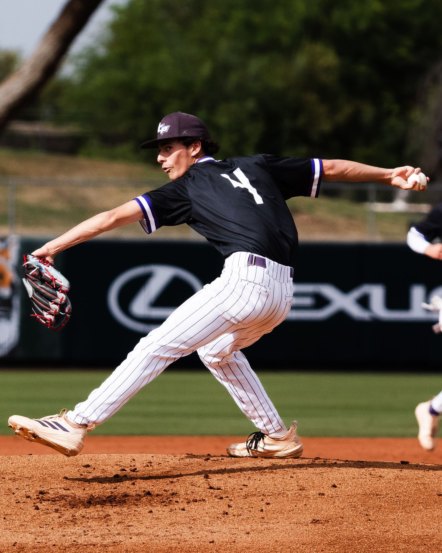 Baseball pitcher in black and white pinstripe uniform mid-throw on mound with Lexus advertisement in background.