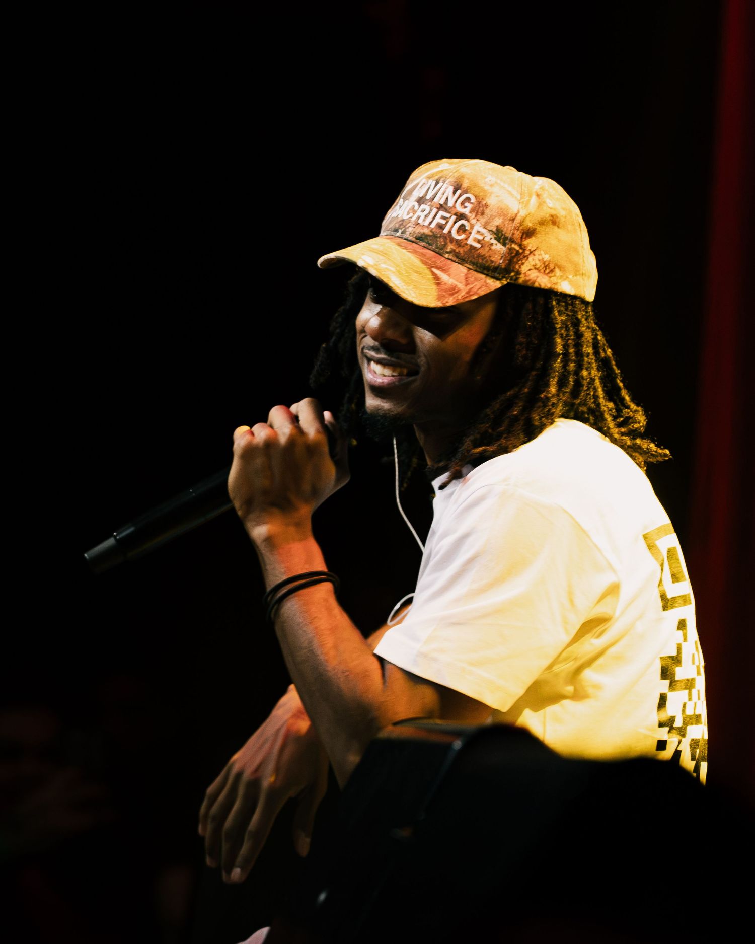 Male performer with dreadlocks and yellow cap singing into microphone on dark stage wearing white tee.