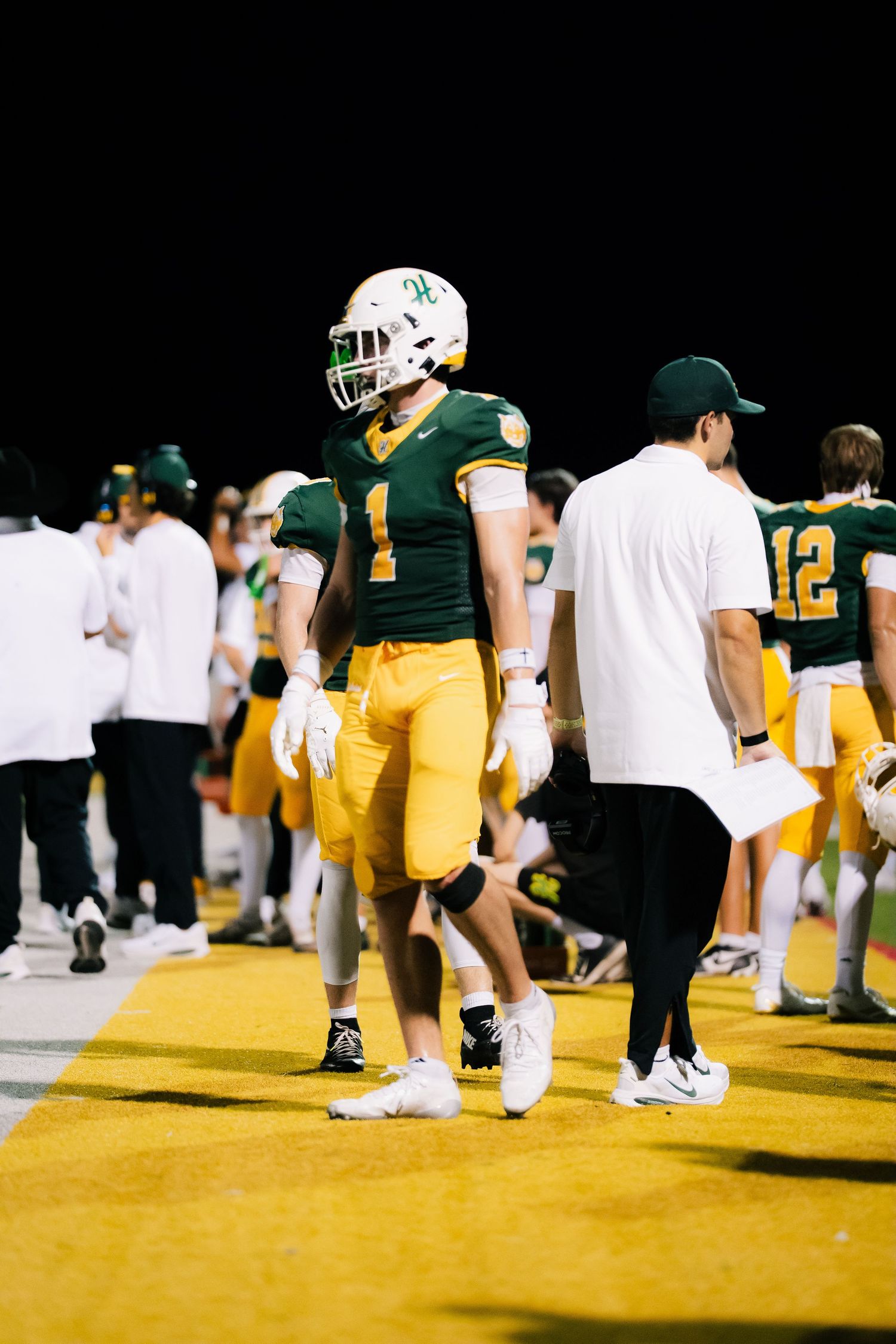 Football player wearing green and gold uniform number 1 stands on sideline during a night game.