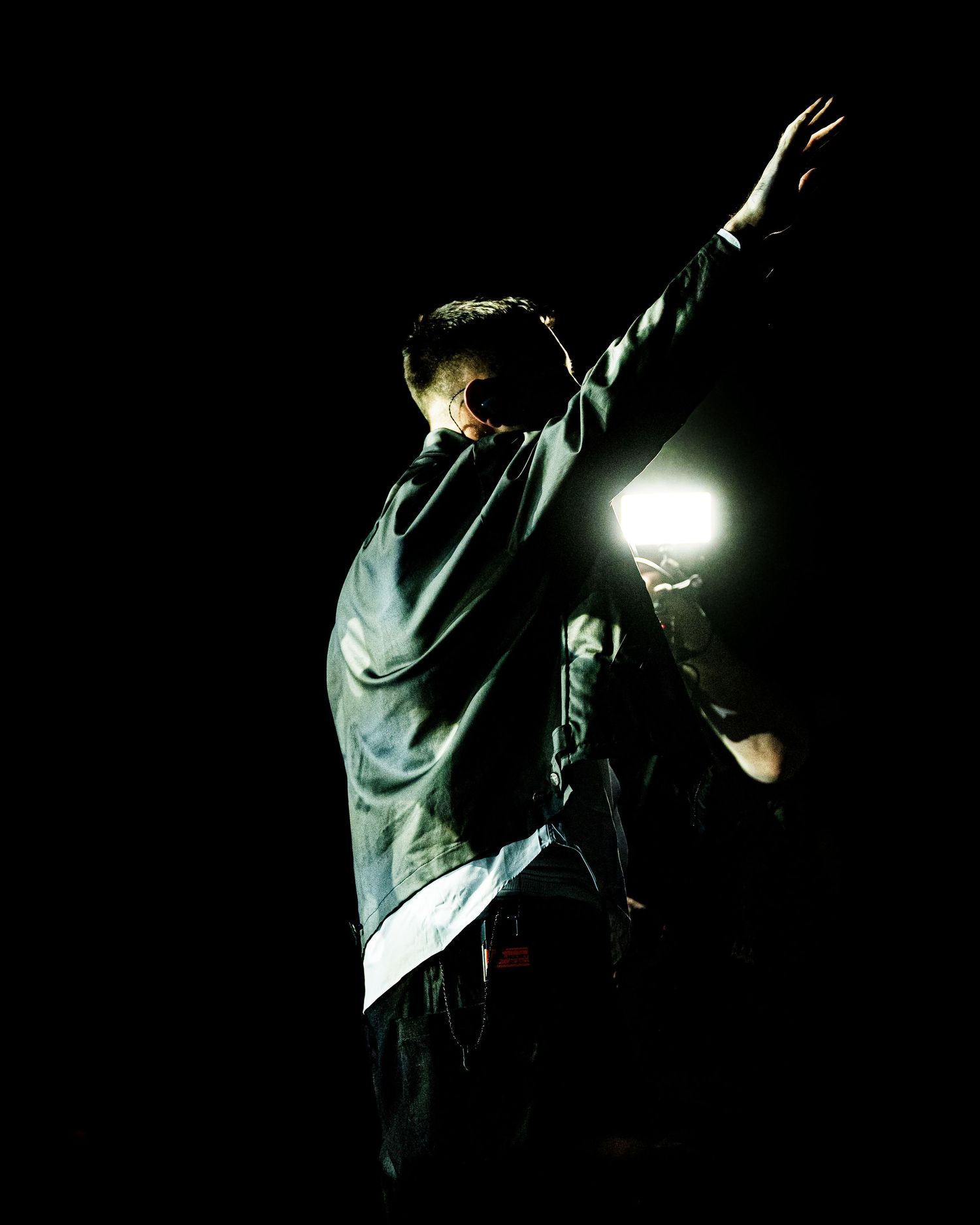 Performer raises arm dramatically on dark stage with bright backlight creating silhouette effect at live concert.