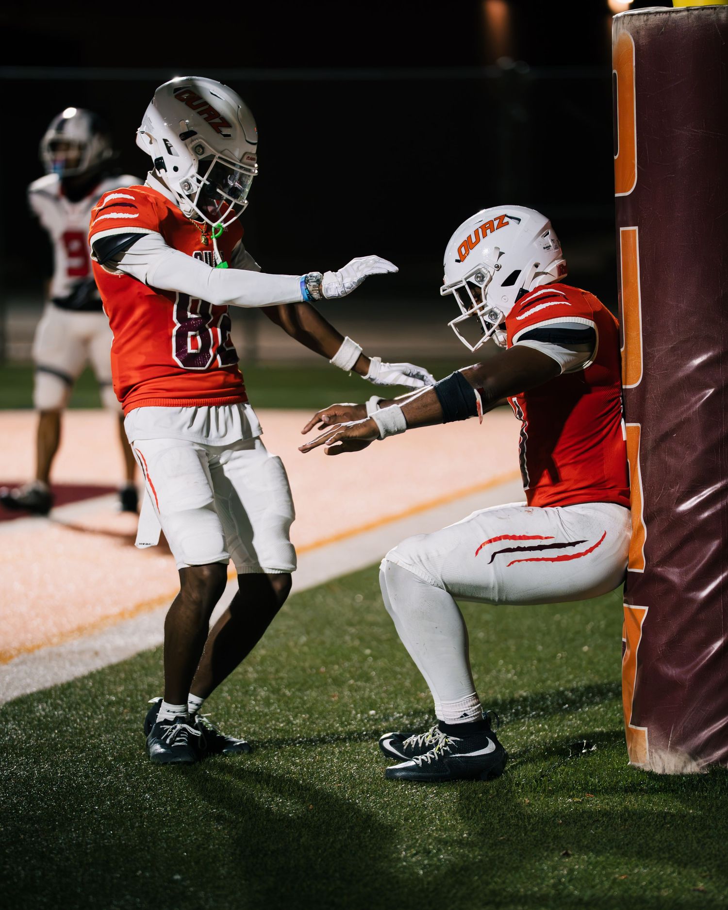 Two football players in orange uniforms celebrate near the end zone during a night game.