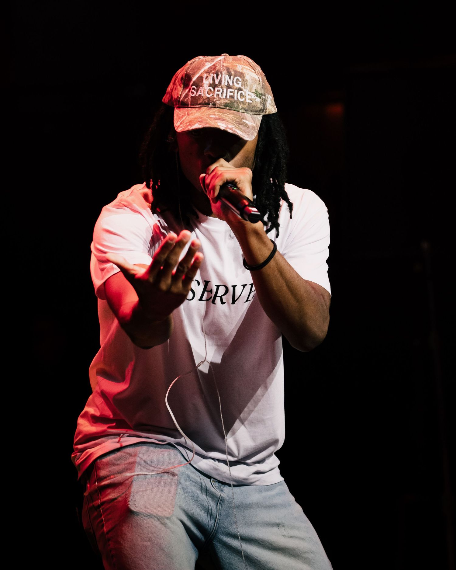 Performer with dreadlocks wearing white SERVE shirt and red cap rapping on stage under dramatic lighting.