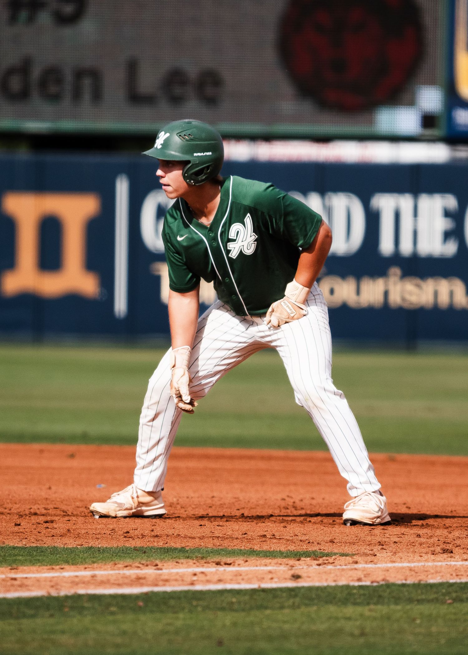 Baseball player in green helmet and pinstripe uniform takes a lead off base on a dirt infield.
