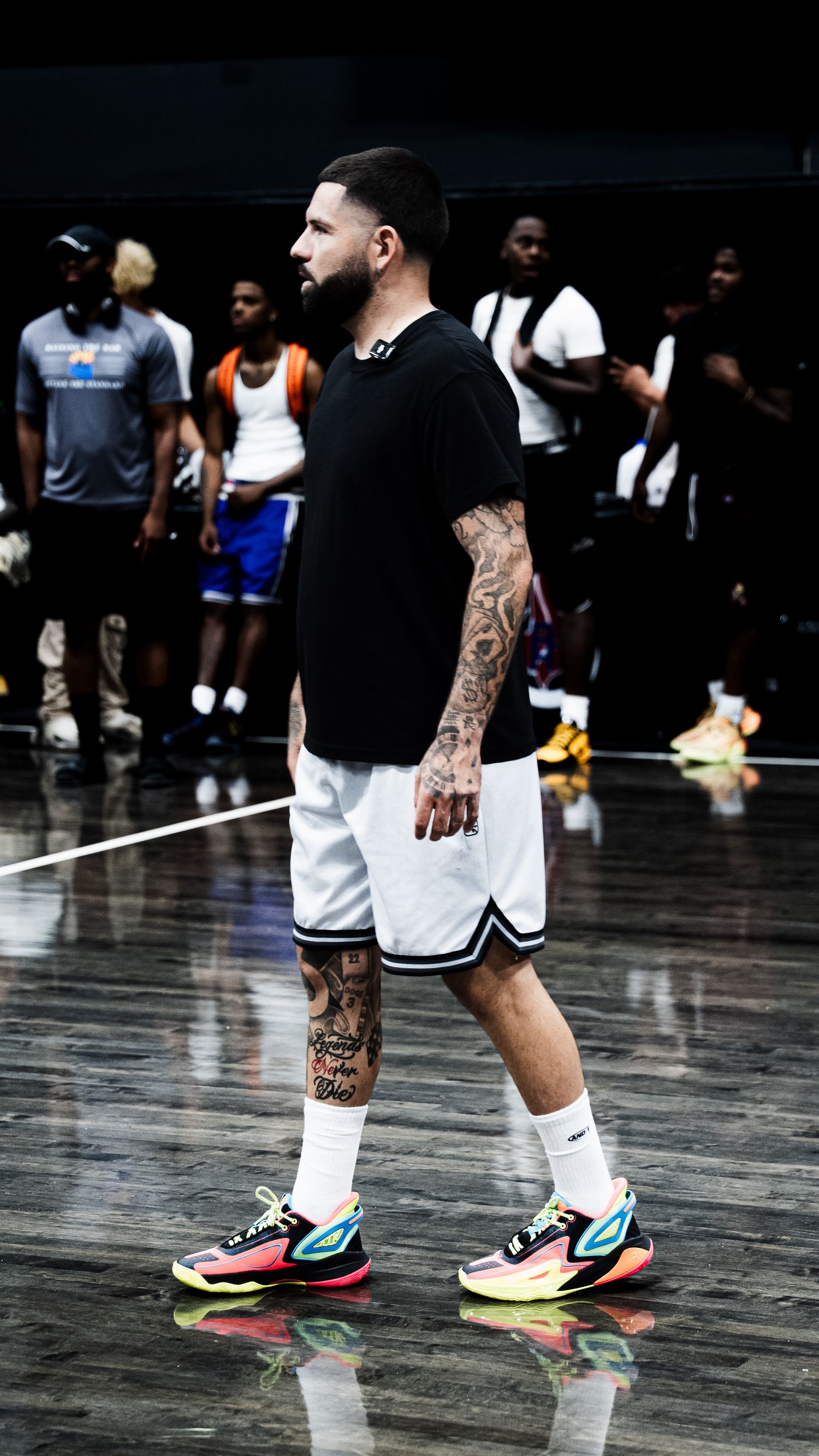 Tattooed male basketball player standing on court in black shirt, white shorts, and multicolor sneakers.