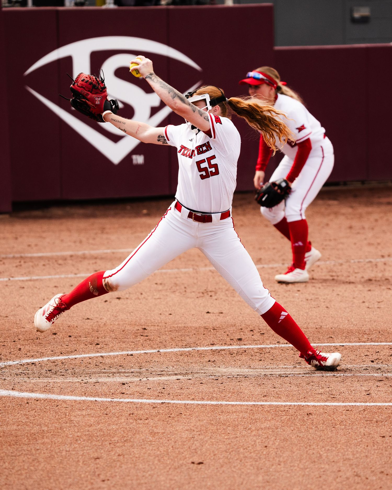 Softball pitcher #55 in white and red uniform delivers a pitch on a dirt field during a college game.