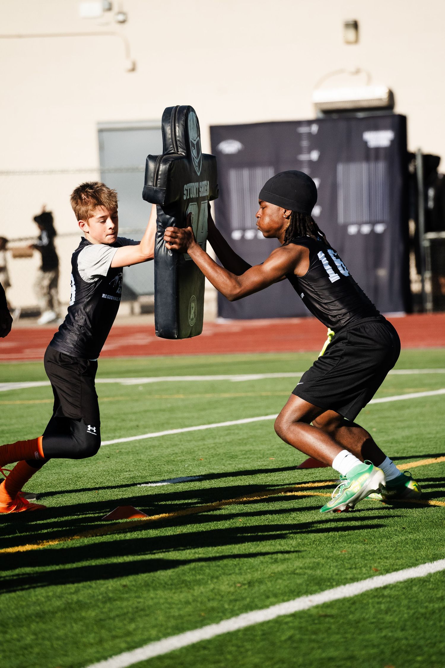 Two youth football players practicing blocking drills with a pad on green turf.