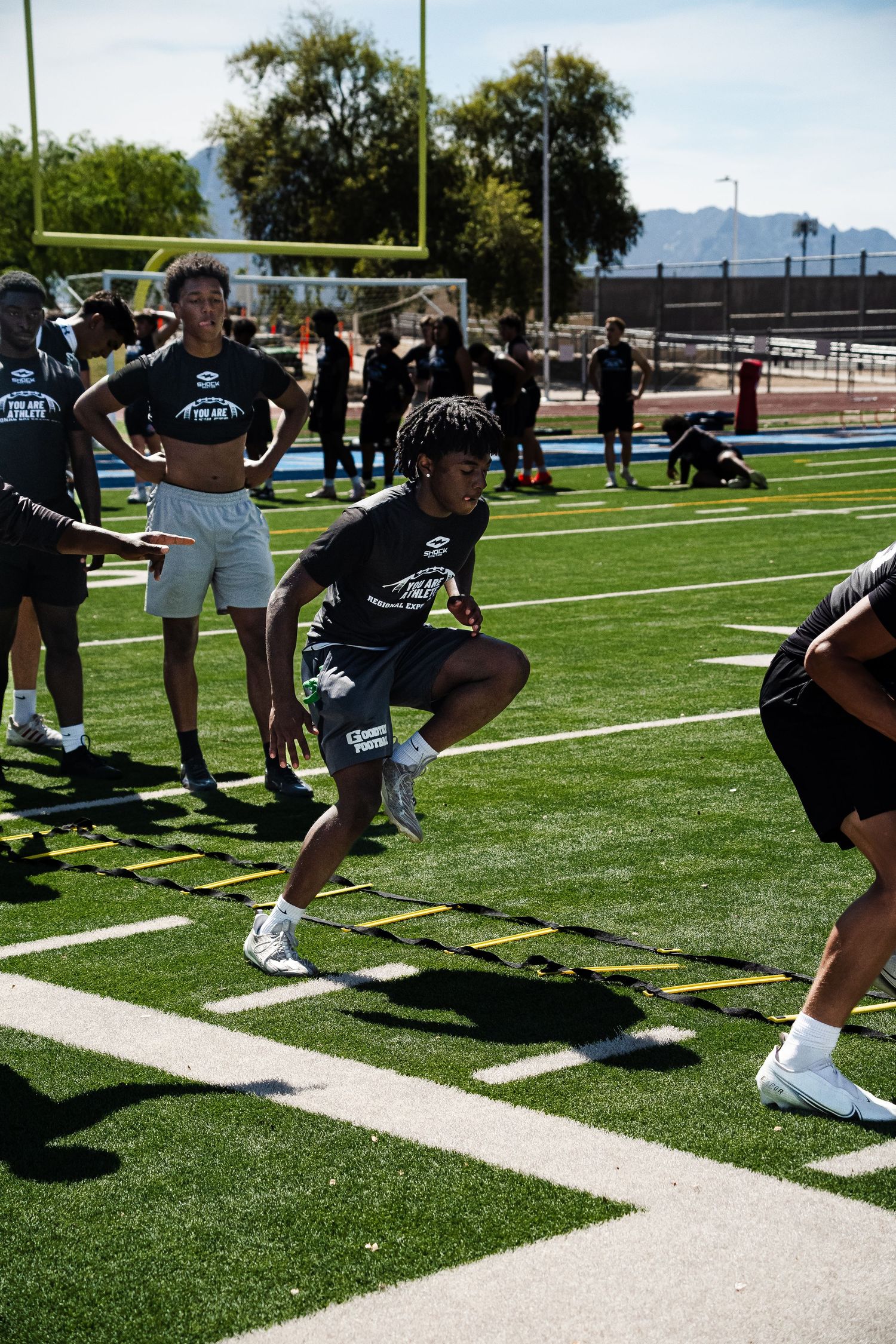 Multiple athletes perform agility ladder drills on sunny outdoor football turf field.