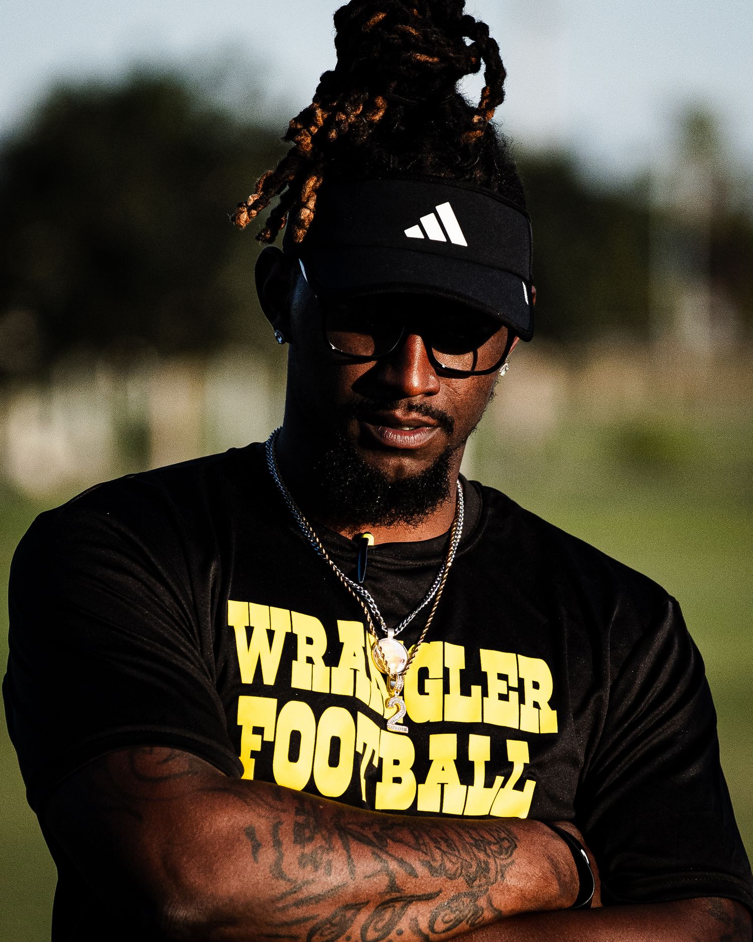 Man in black Adidas cap and Wrangler Football shirt with dreadlocks, tattoos, and gold chains poses outdoors.