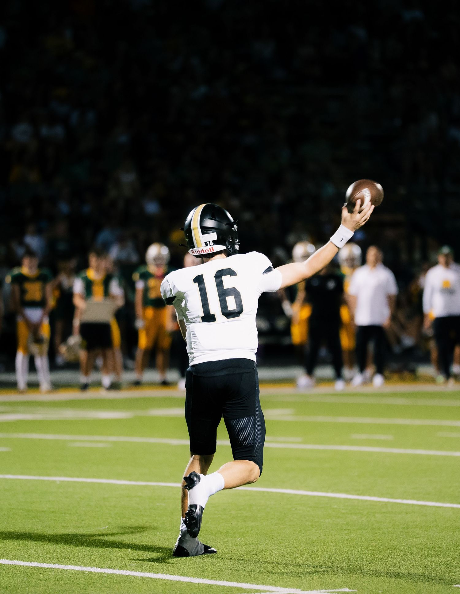 High school football player #16 in white uniform throws a pass on a lit field during a night game.