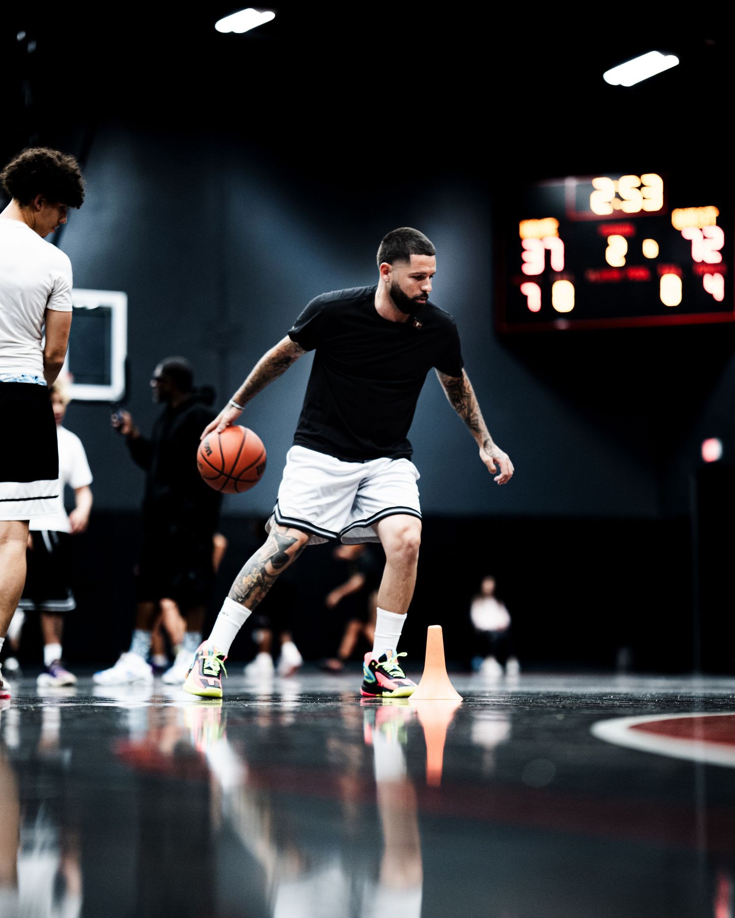 Tattooed basketball player dribbling on indoor court during practice session with scoreboard in background.