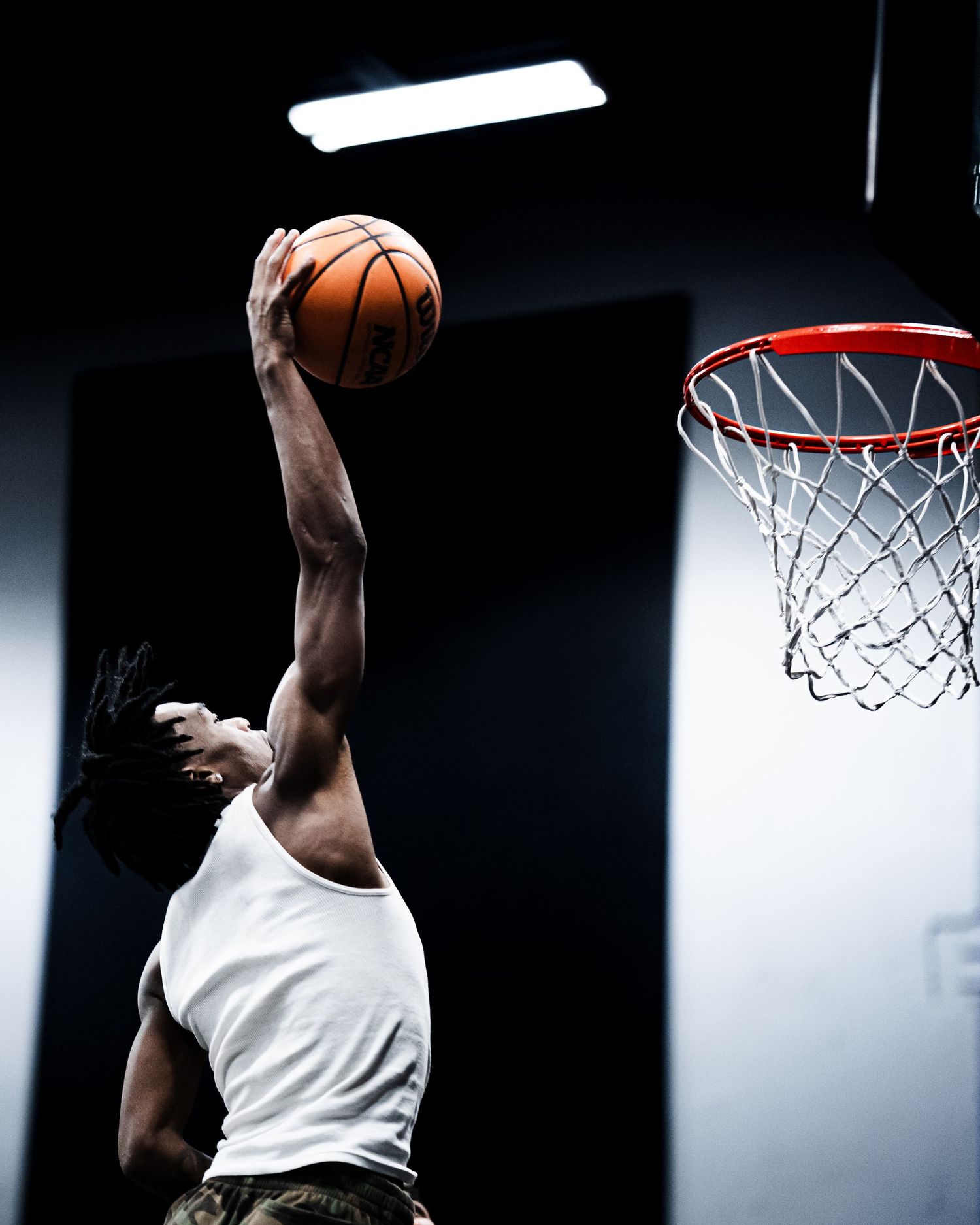 A basketball player leaps to dunk an orange ball into a red-rimmed hoop in a dimly lit indoor court.