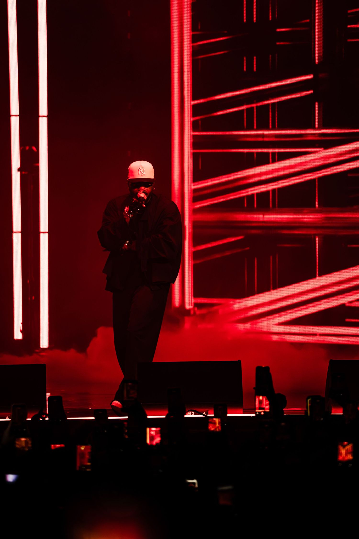 A performer in a white cap and dark clothing sings into a microphone on a stage lit by red lights and glowing metal scaffolding.