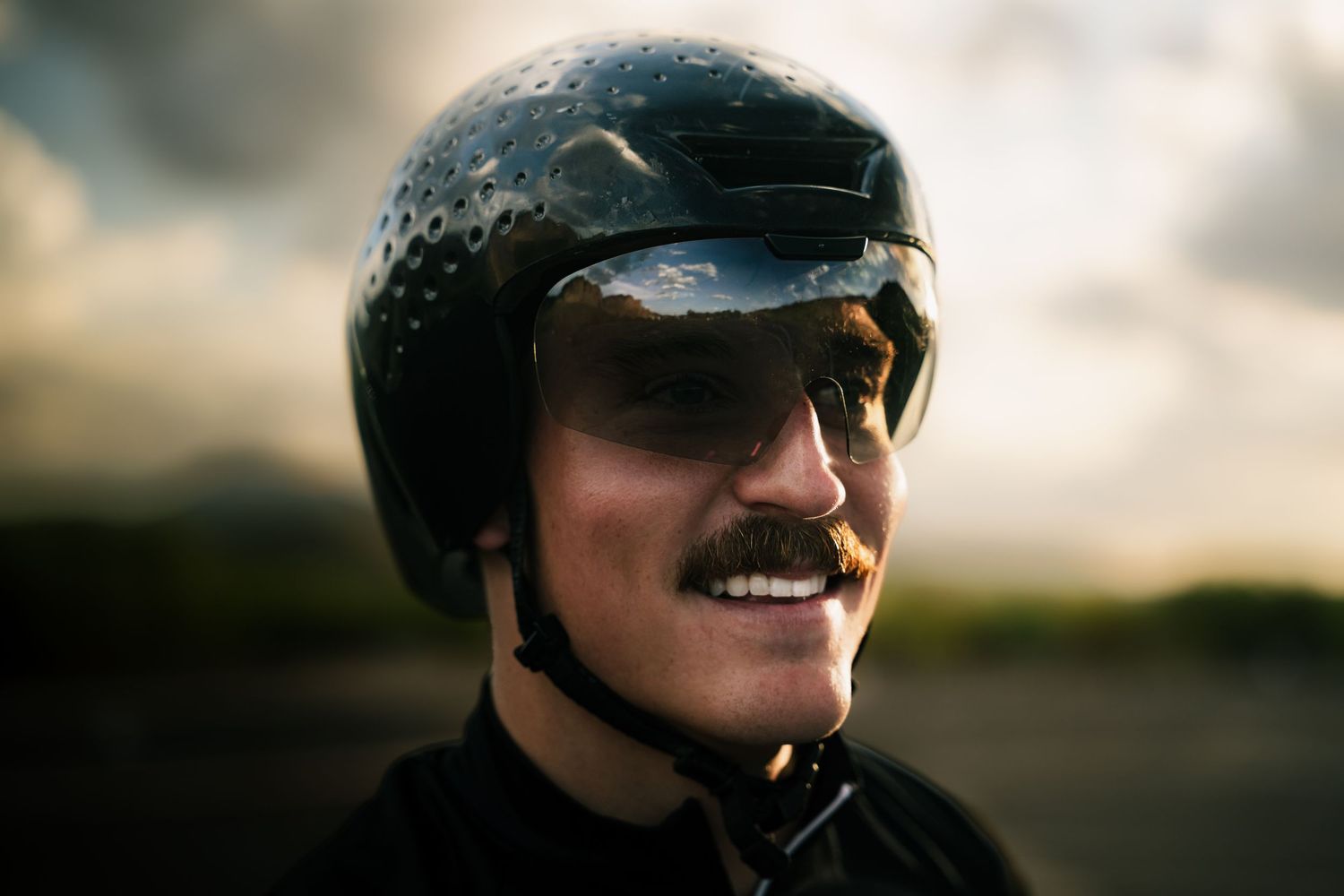 Smiling man wearing a black motorcycle helmet and goggles outdoors under a dramatic cloudy sky.