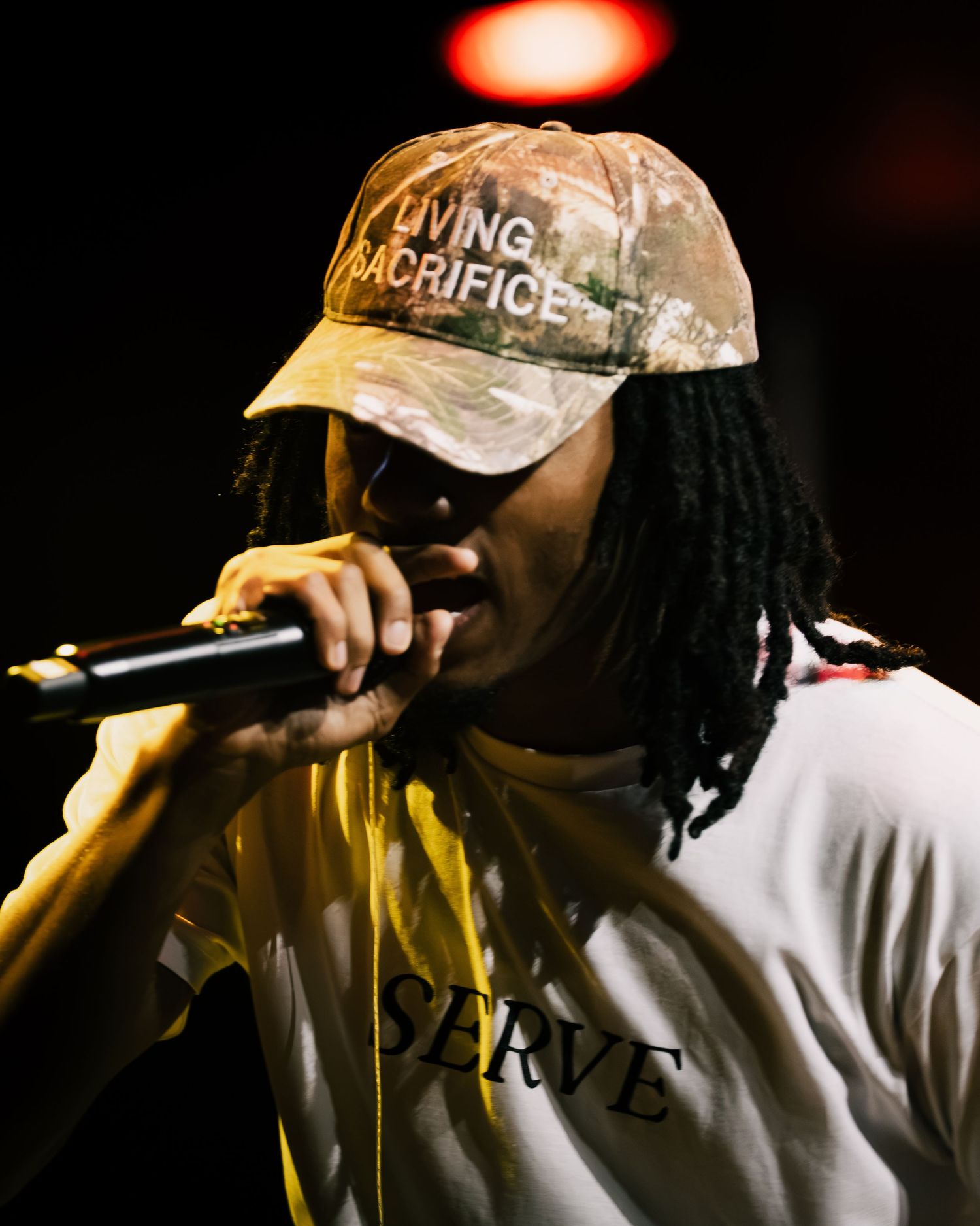 Young male rapper performing on stage wearing a camo cap and white SERVE t-shirt while holding a microphone.