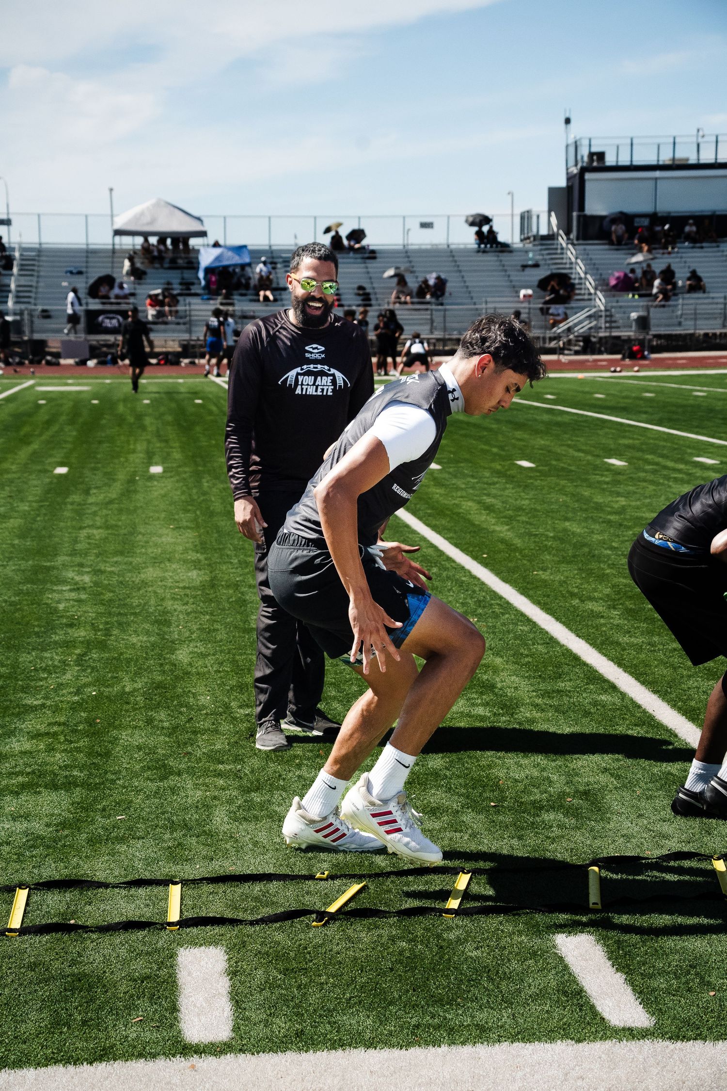 Coach in green sunglasses watches athlete perform agility ladder drill on football field.