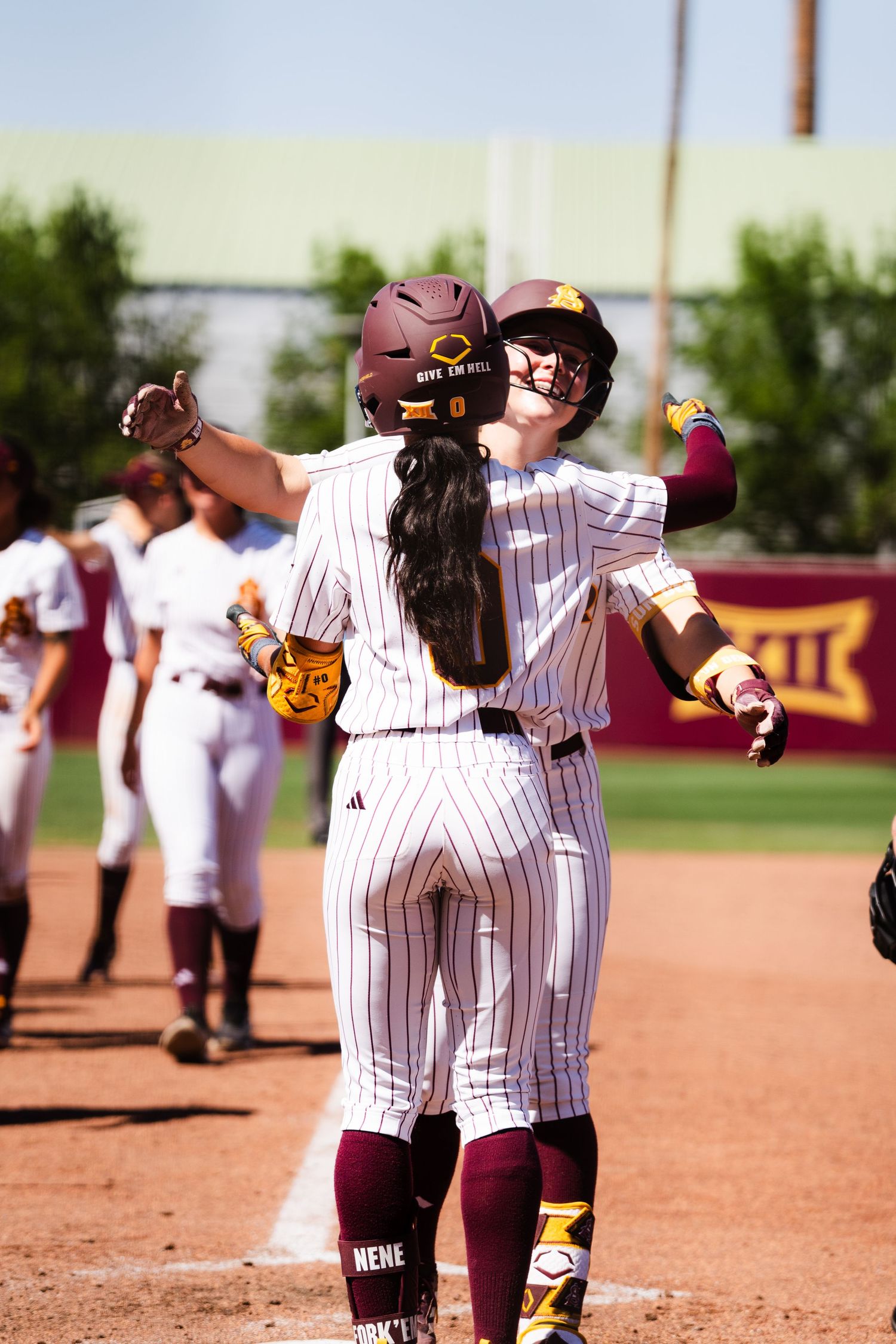 Two female college softball players in maroon pinstripe uniforms embrace in celebration after a home run.